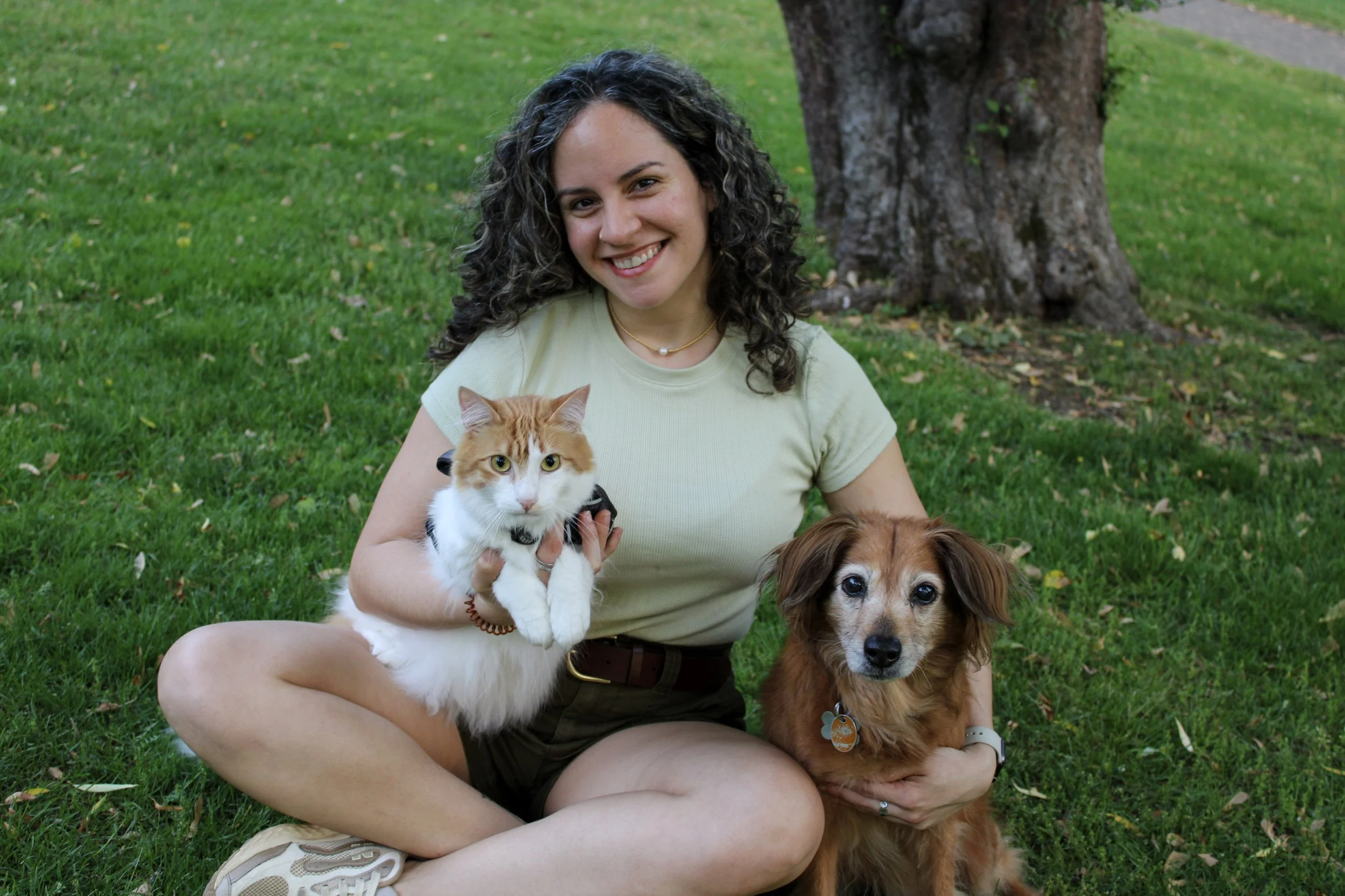 A woman pet sitter smiling and sitting on grass outdoors, holding a white and orange cat; a brown dog sitting nearby, in front of a large tree.