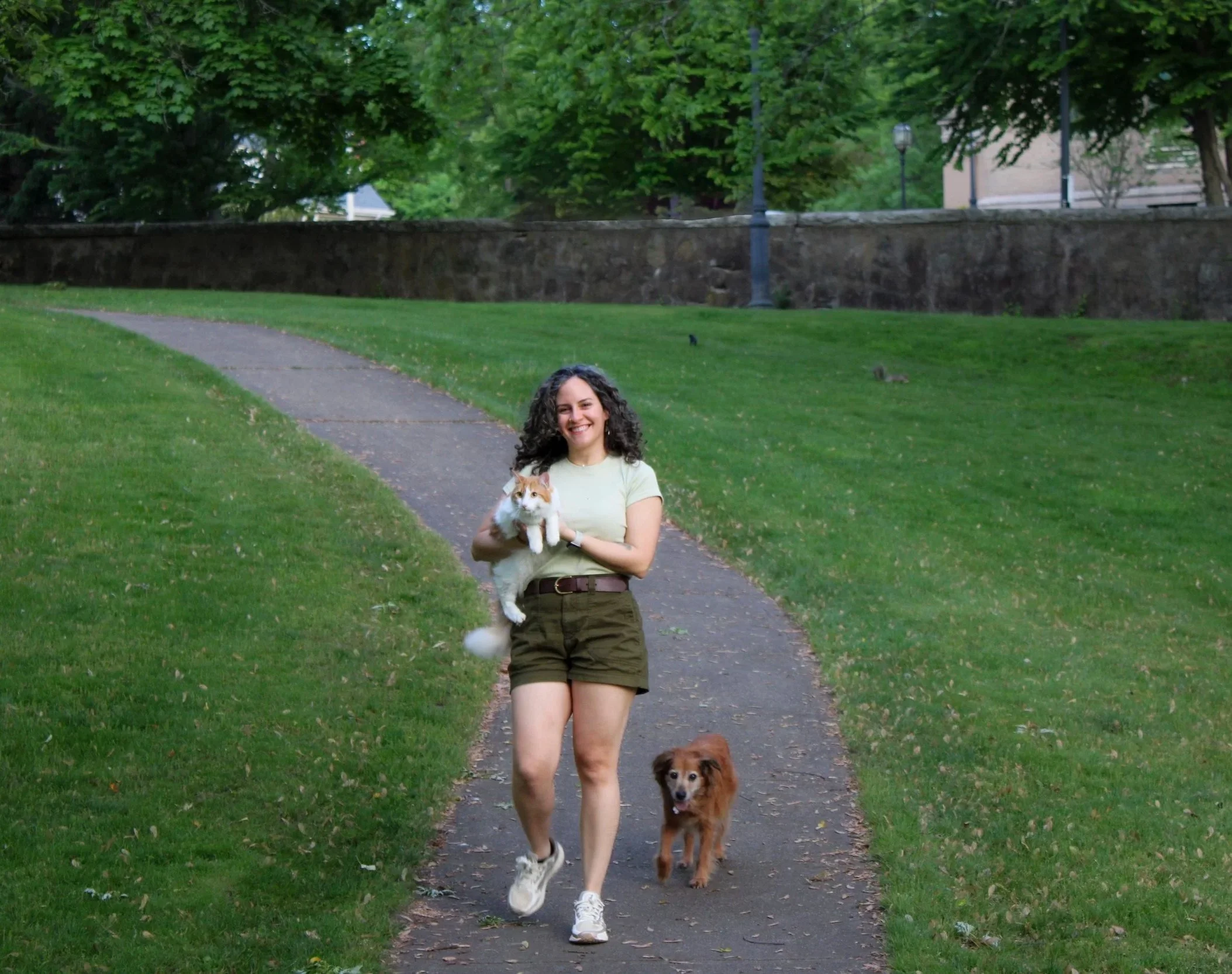 A woman pet sitter smiling and walking outdoors on a sidewalk, holding a white and orange cat, with a dog walking beside her in a park with green grass and trees.