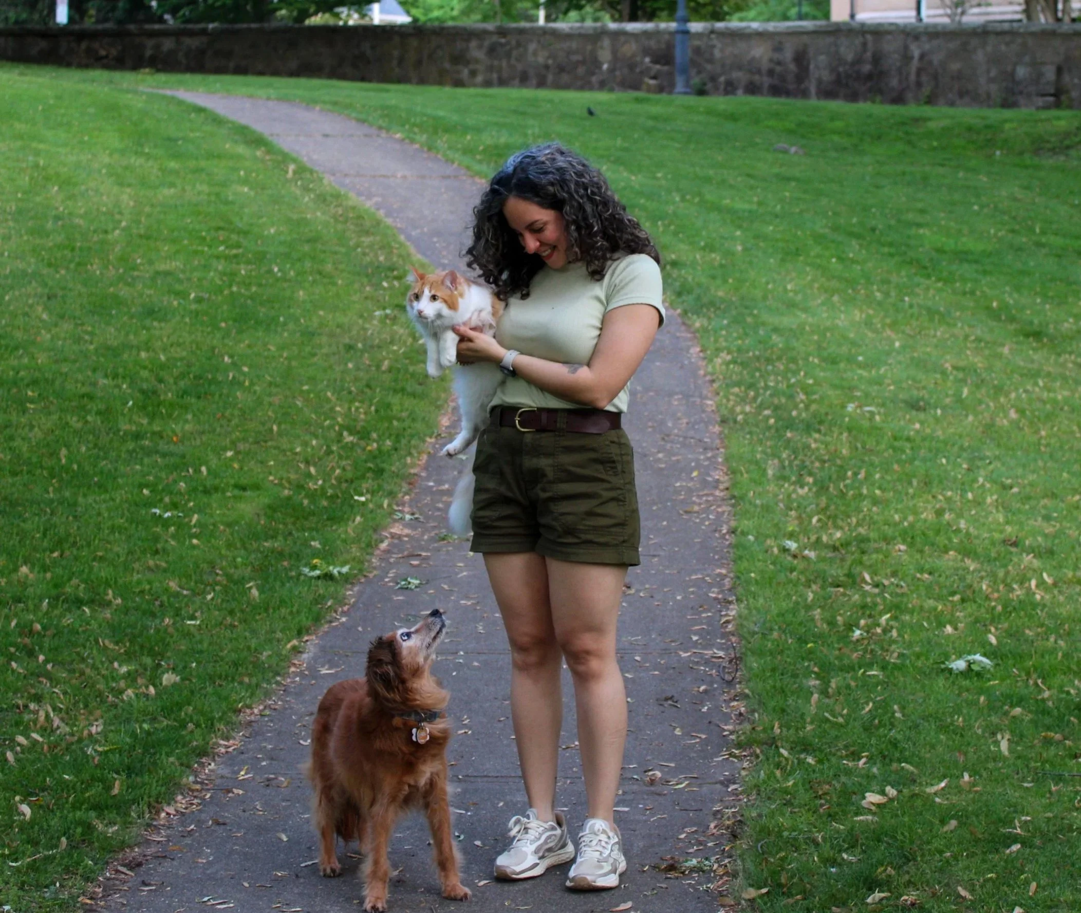 A woman pet sitter smiling while holding a small orange and white, and a brown dog by her side looking at her at the park