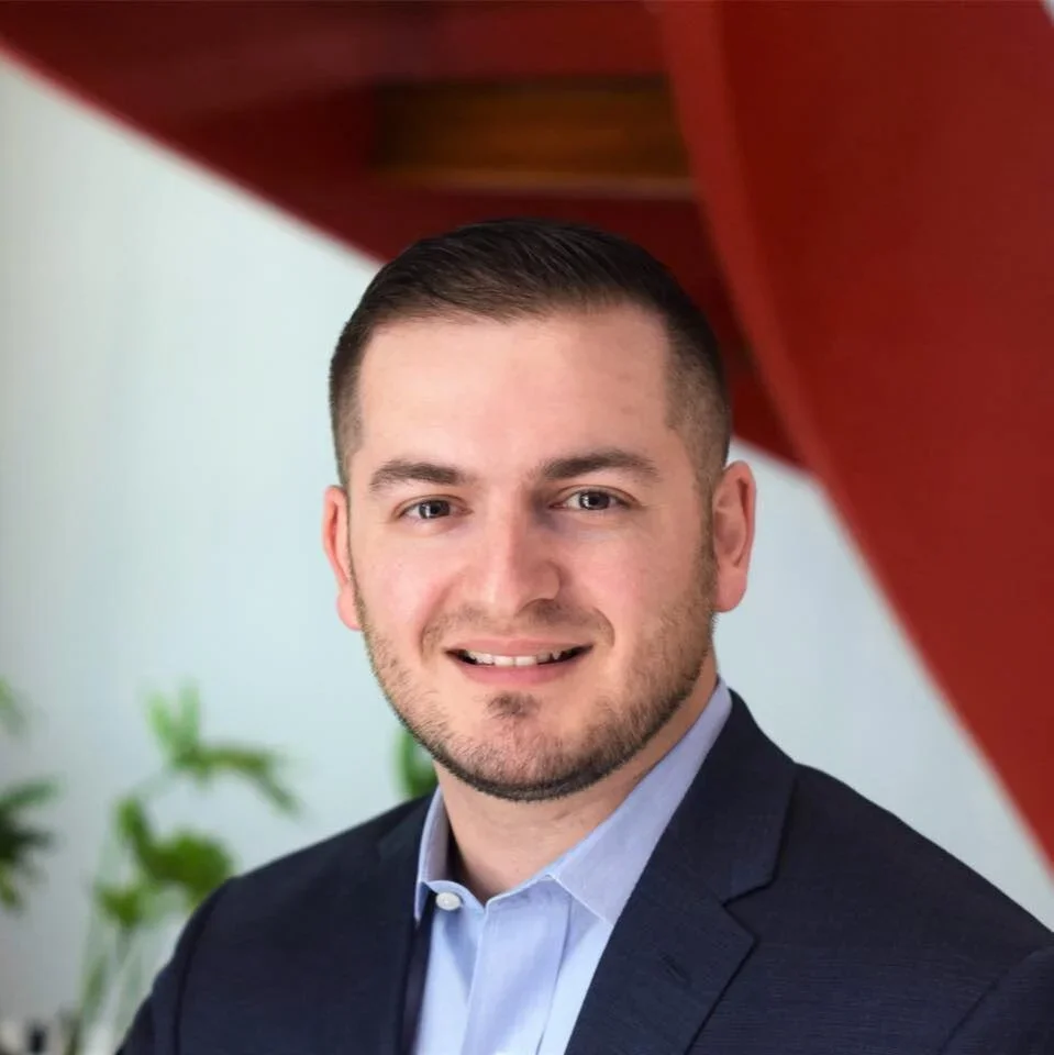 A young man (Jake Sarfati) with short dark hair, a beard, and wearing a navy suit jacket and light blue collared shirt, smiling at the camera. The background includes a plant and a red structure.