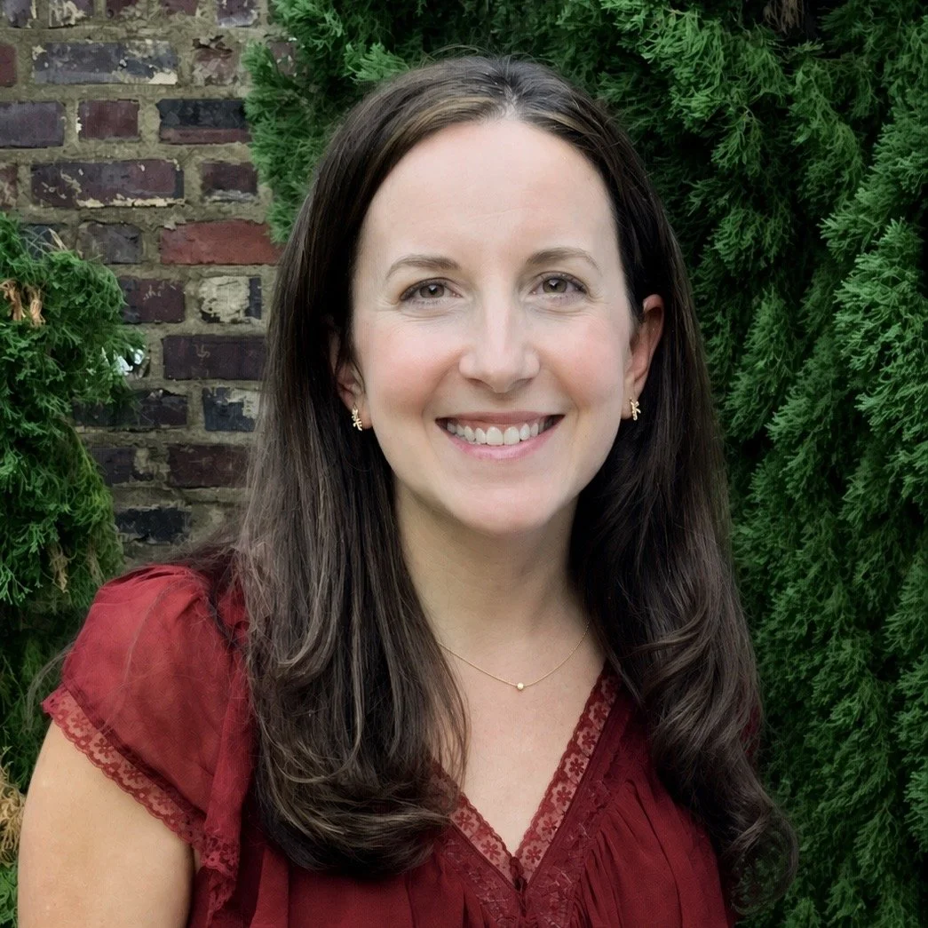 A woman with long dark hair smiling in front of a brick wall and green foliage.