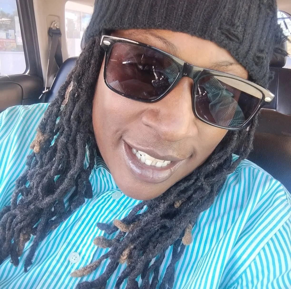 A person with dreadlocks wearing sunglasses, a gray beanie, and a turquoise striped shirt, smiling inside a vehicle.