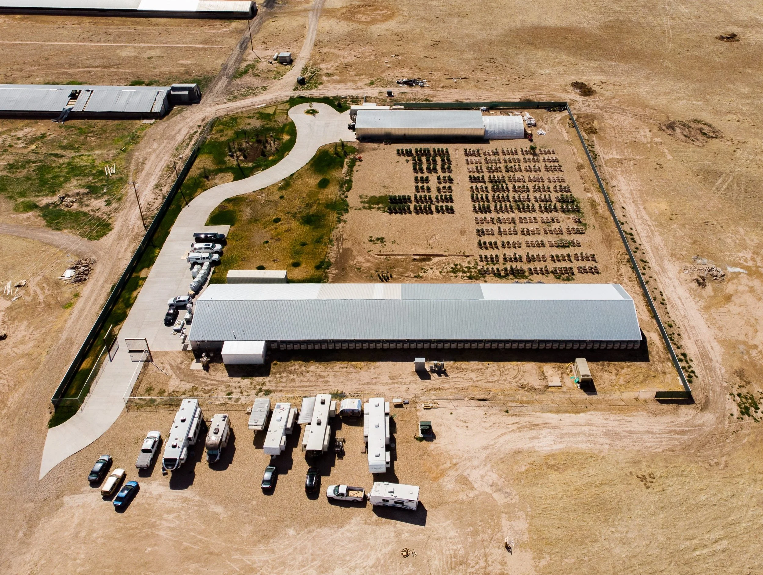 Aerial view of a construction or storage site with several parked vehicles, storage units, and a fenced-in area with organized rows of plants, set in a barren landscape with dirt and minimal vegetation.