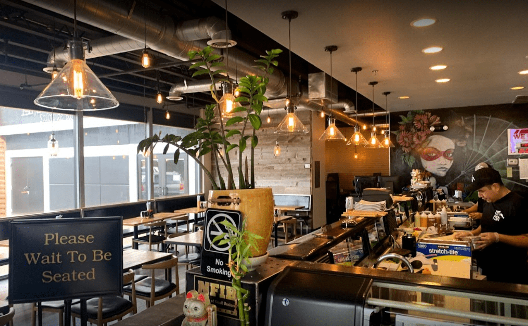 Interior of a modern restaurant with wooden tables and chairs, a large potted plant, and a colorful mural of a woman's face with glasses on the wall. Pendant lights hang from the ceiling, and a person working at the counter is visible.