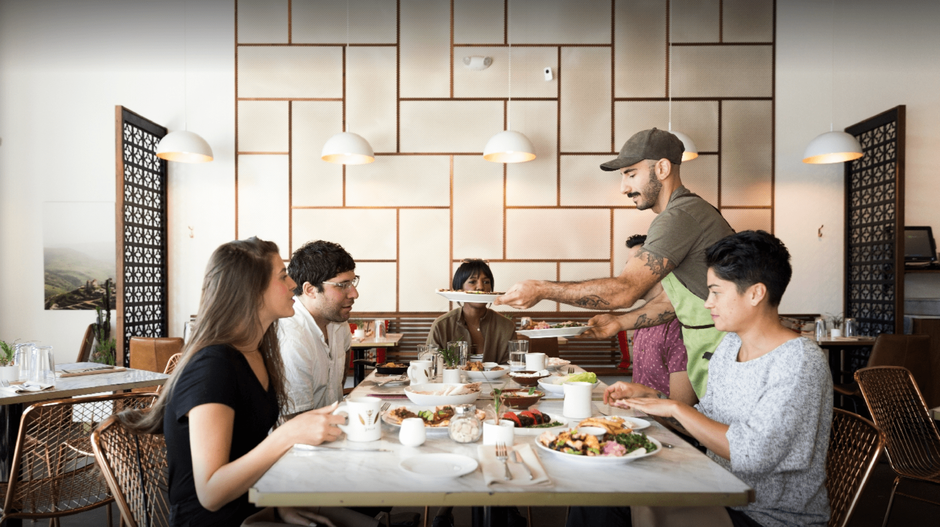 A group of five diverse people sitting around a dining table in a restaurant, being served by a waiter. The table is filled with plates of food, drinks, and tableware, with a modern interior featuring wooden wall accents and hanging lights.
