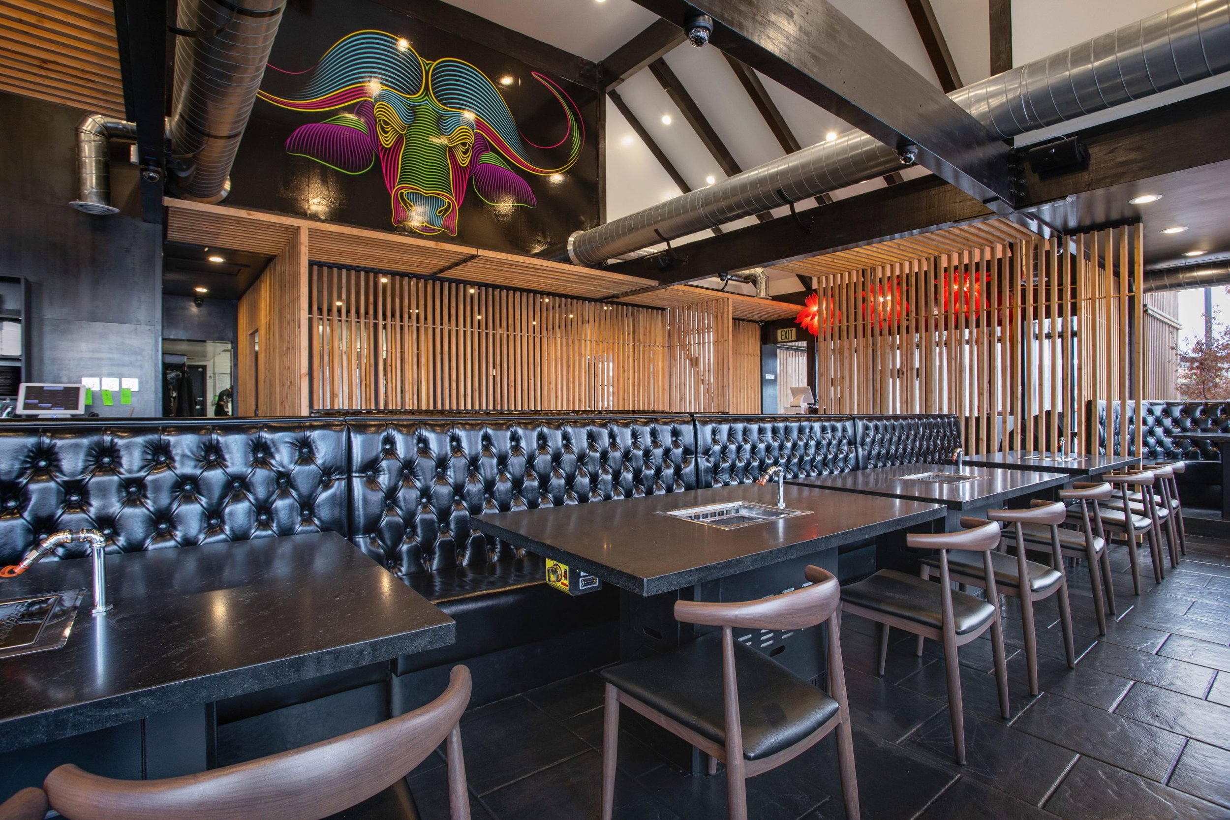 Interior of a modern restaurant with black tables and brown chairs, wood paneling, exposed ductwork, and a colorful neon bull head art on the wall.