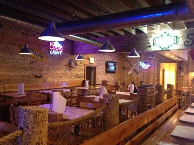 Empty restaurant interior with wooden tables, chairs, and cozy lighting, featuring neon signs and nautical decor.