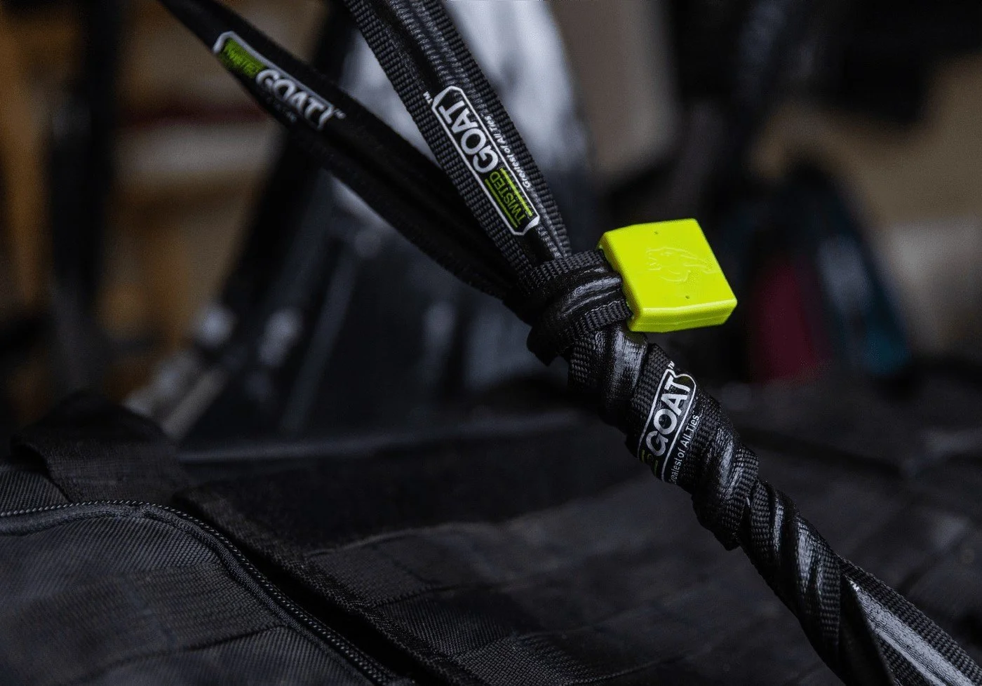 Close-up of a black protective strap with labels reading 'G.O.A.T' and 'GORE', secured with a neon green lock, attached to a black bag or equipment.