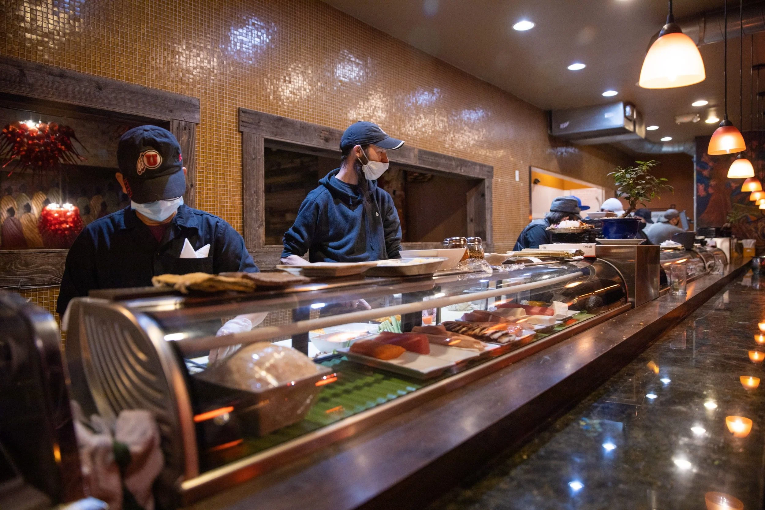 Restaurant workers wearing masks behind a sushi bar with various sushi dishes on display.