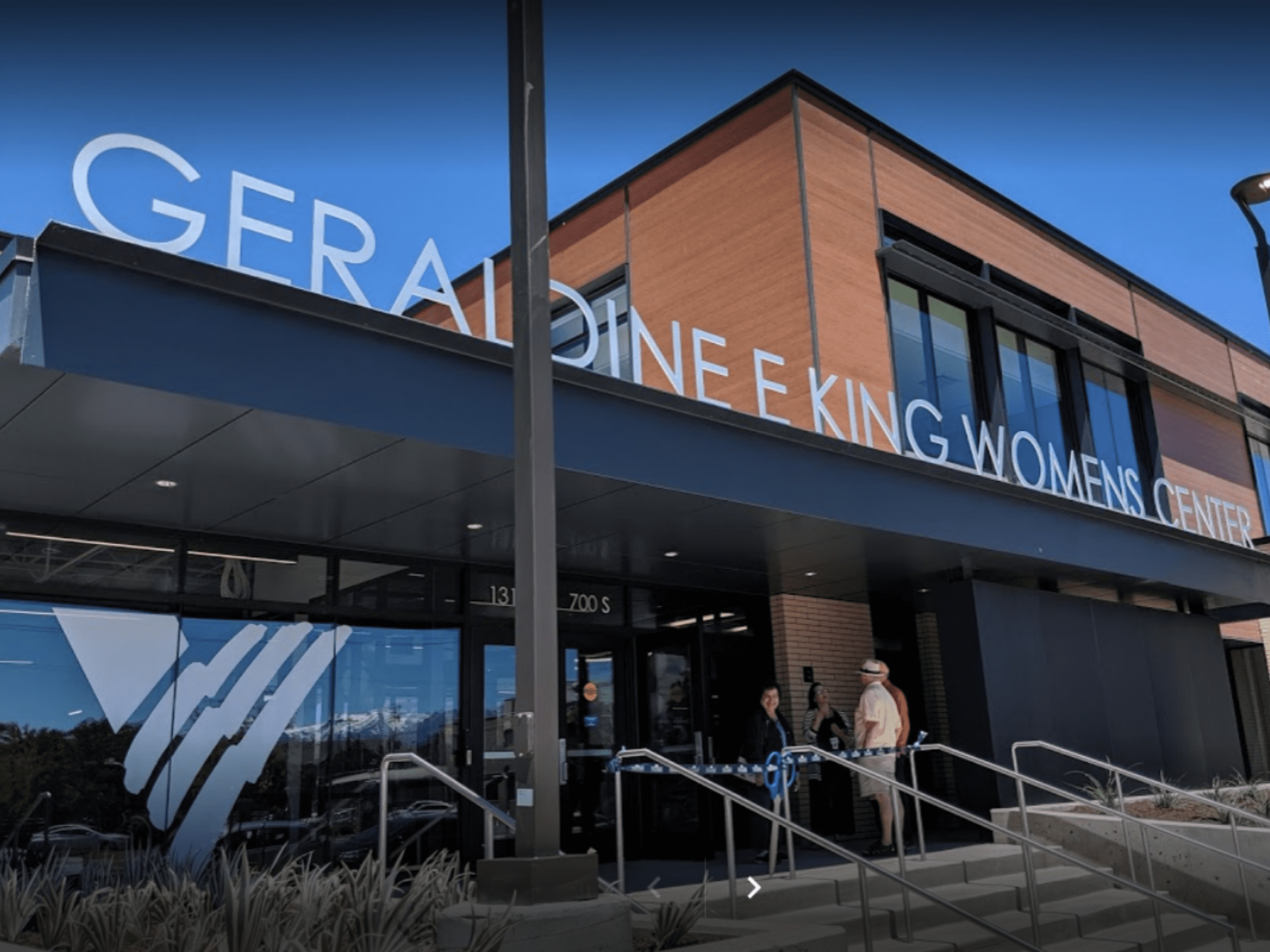 Exterior view of a modern building with a large sign reading 'Geriatric Women's Center' on the roof, with a blue sky in the background, and a few people standing outside near the entrance.