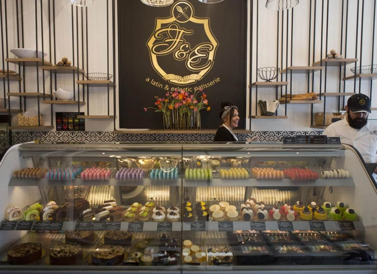Display case filled with colorful macarons, petit fours, and cakes in a patisserie. Two employees behind the counter, one woman smiling and one man wearing a chef's coat and cap. A large black board with a logo reading 'F&E: a Latin & Electric Patisserie' and a flower arrangement on a shelf in the background.