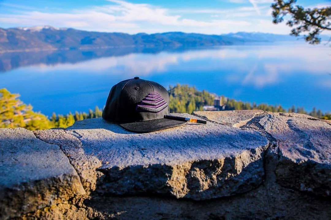 A black baseball cap with purple stripes and a brush resting on a stone wall, overlooking a calm lake and forested hills in the distance.