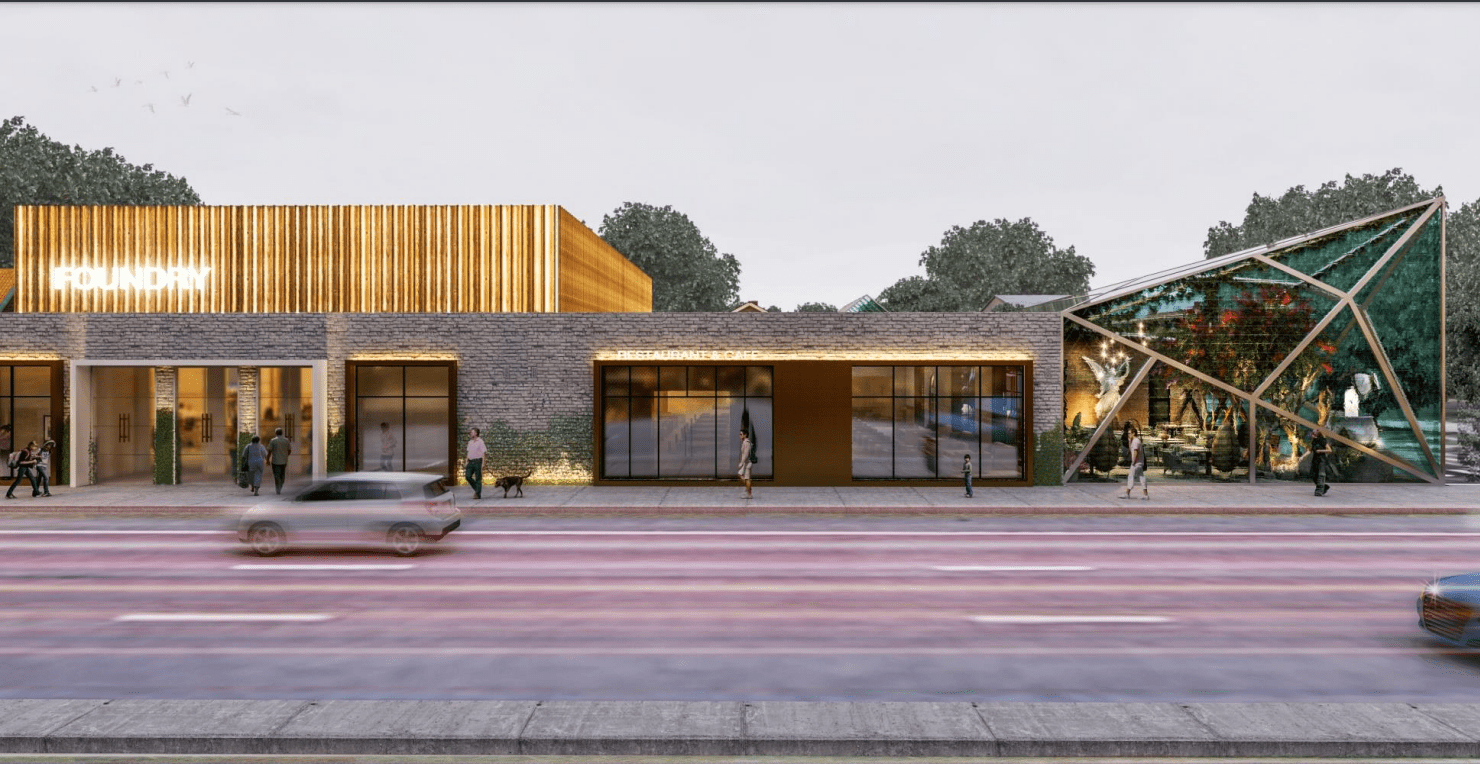 A modern building with a mix of brick, wood, and glass exterior, featuring a brightly lit sign that reads 'FOUNDRY'. There is a glass enclosure on the right with people inside, and pedestrians walking on the sidewalk in front of the building. Blurred cars are passing by on the street.