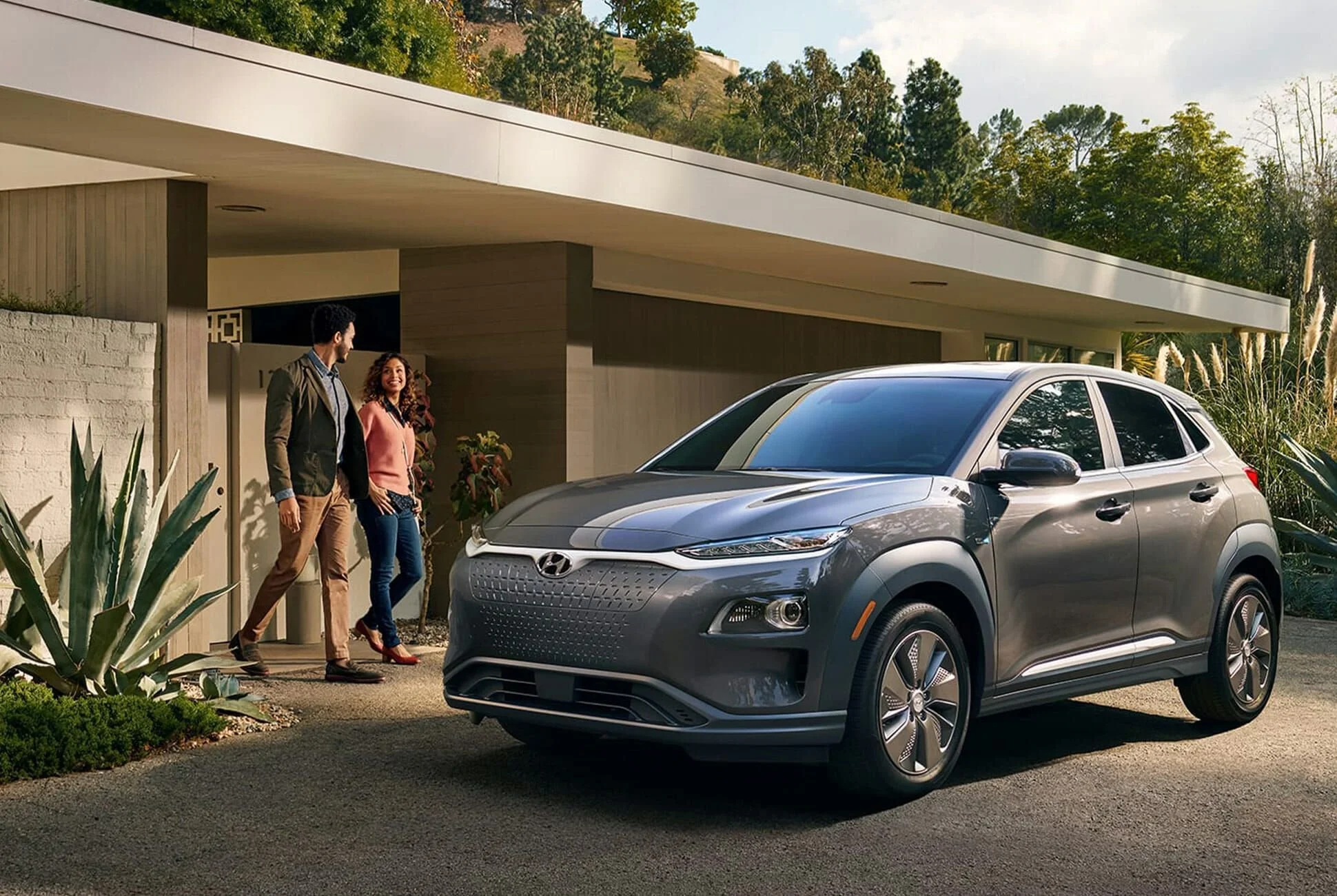 Two people, a man and a woman, standing near a modern house with a gray Hyundai Kona electric car parked in front. The house has a flat roof, wood paneling, and surrounded by greenery, with a backdrop of trees and hills.
