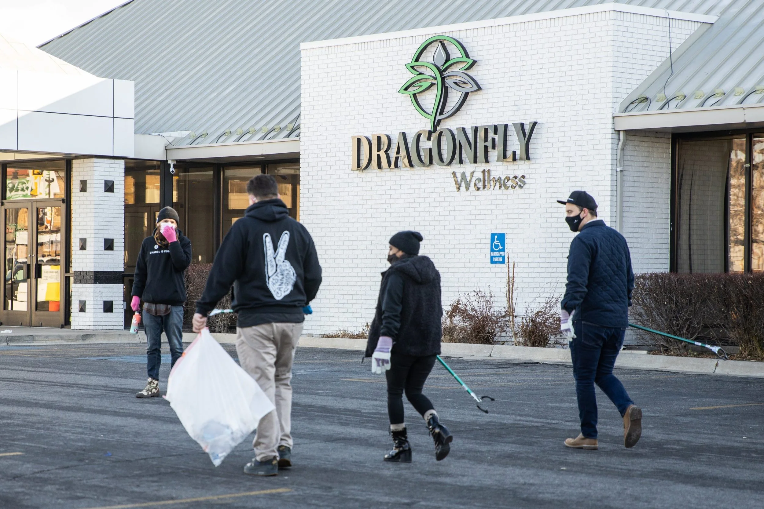 People walking out of a wellness center called Dragonfly Wellness, with three people wearing masks and gloves, some holding cleaning tools, and a large white-brick building with a logo of a dragonfly with leaves.