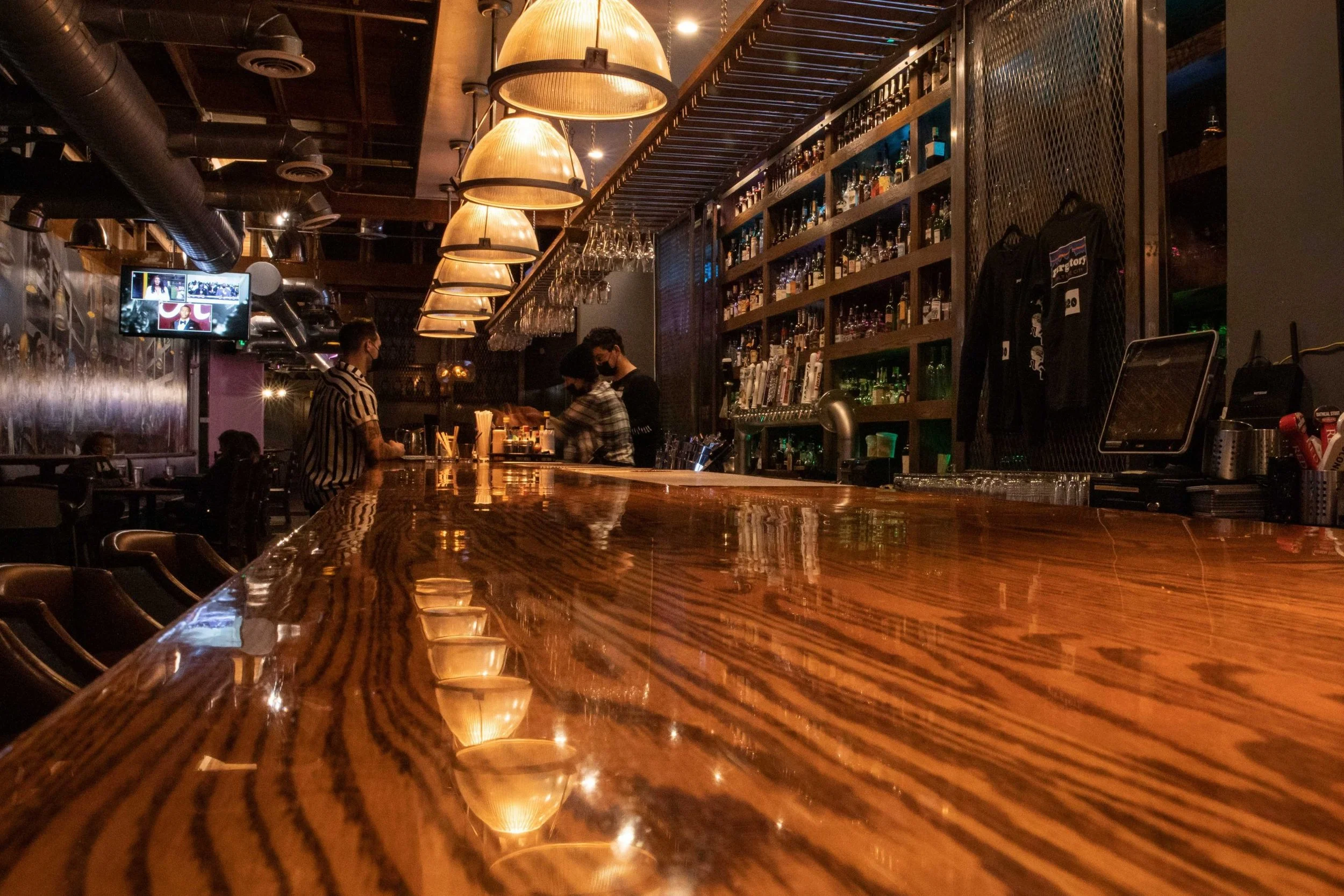 Interior of a dimly lit bar with a polished wooden counter, hanging lamps, and shelves filled with liquor bottles. Three people are standing at the bar, two of them are bartenders or servers. A TV screen is on the wall with multiple images displayed.