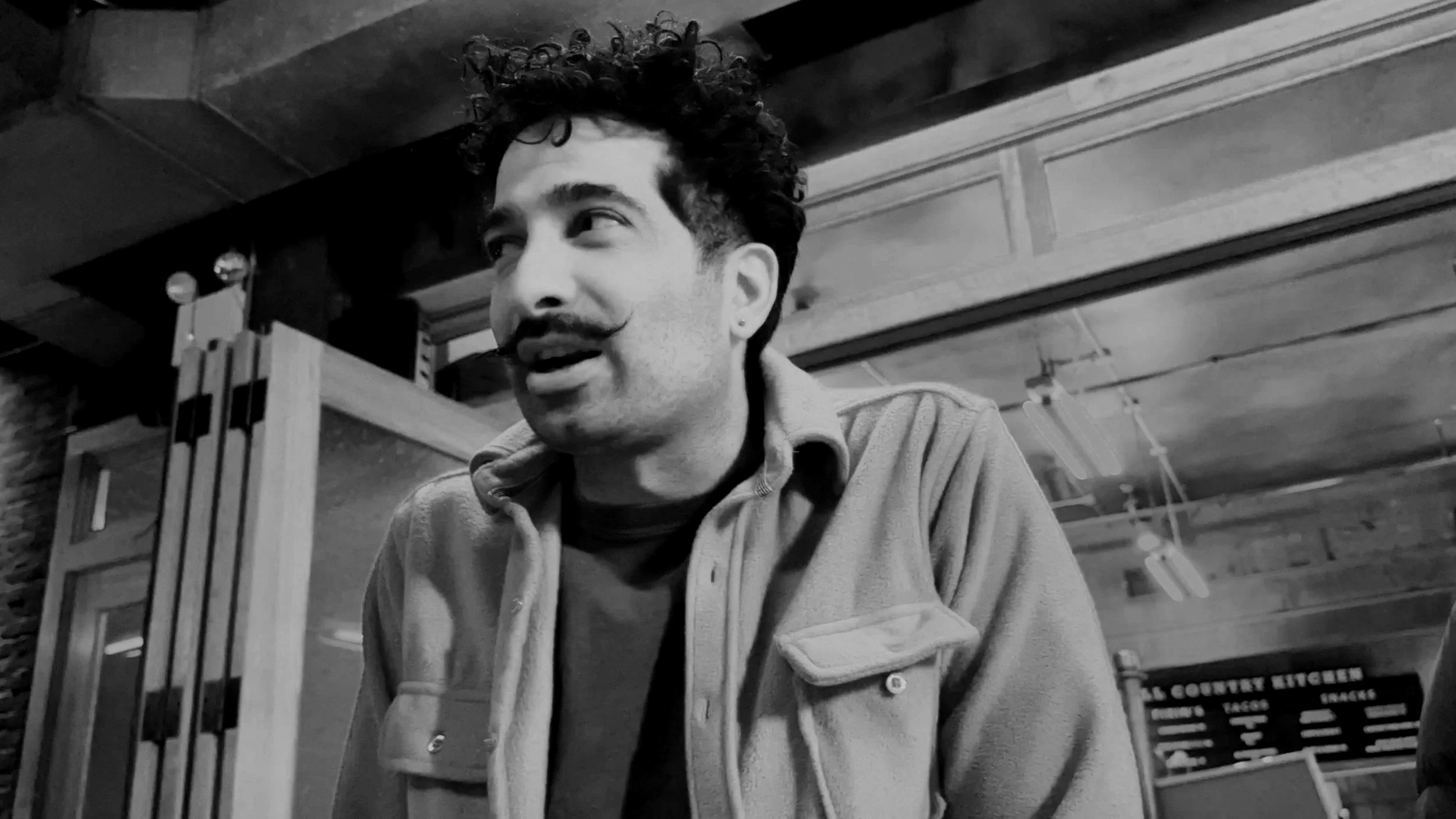 A man with curly hair and a mustache seated in an indoor space.