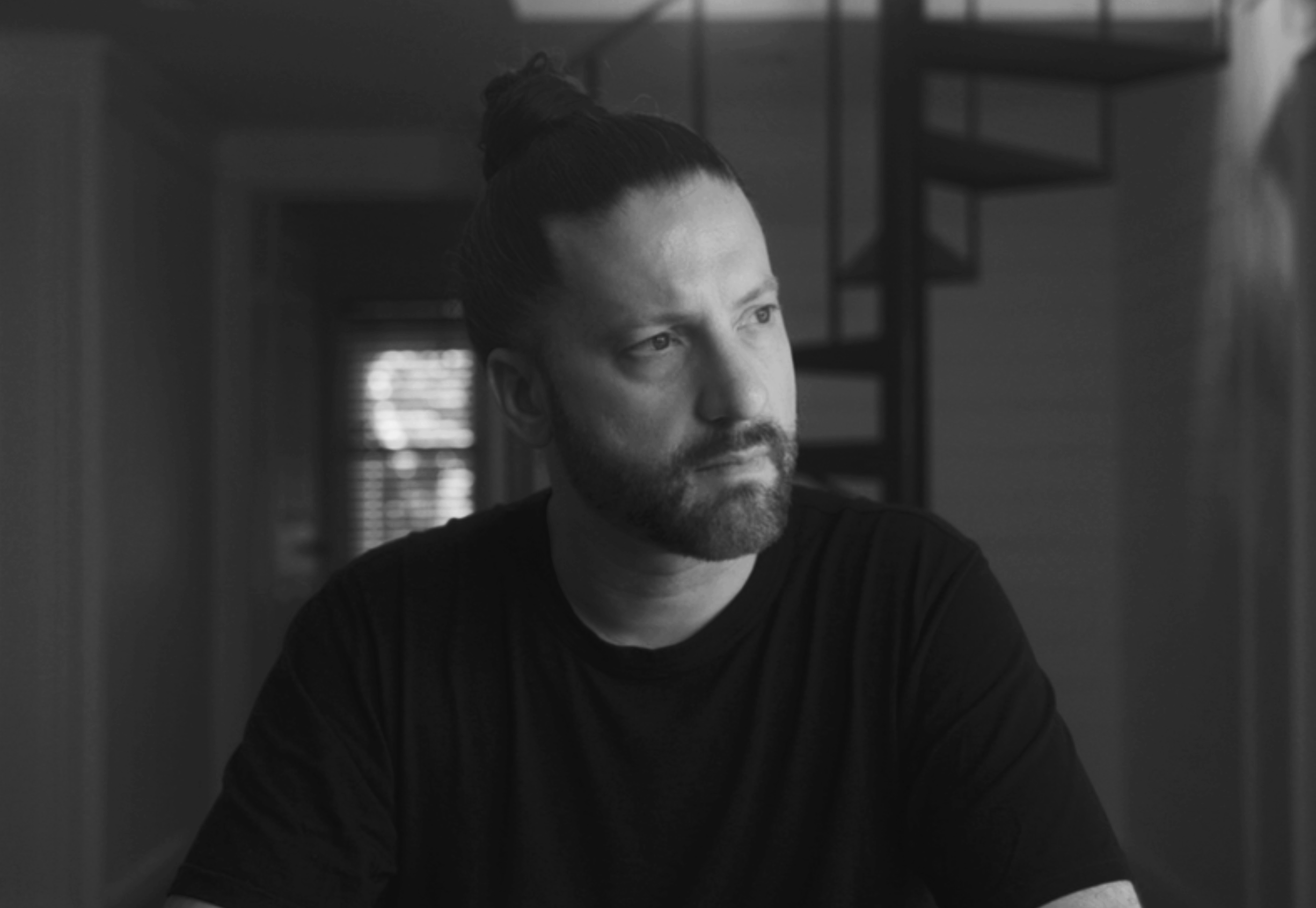A black and white photo of a man with a beard and hair tied up in a bun sitting indoors, with a staircase in the background.