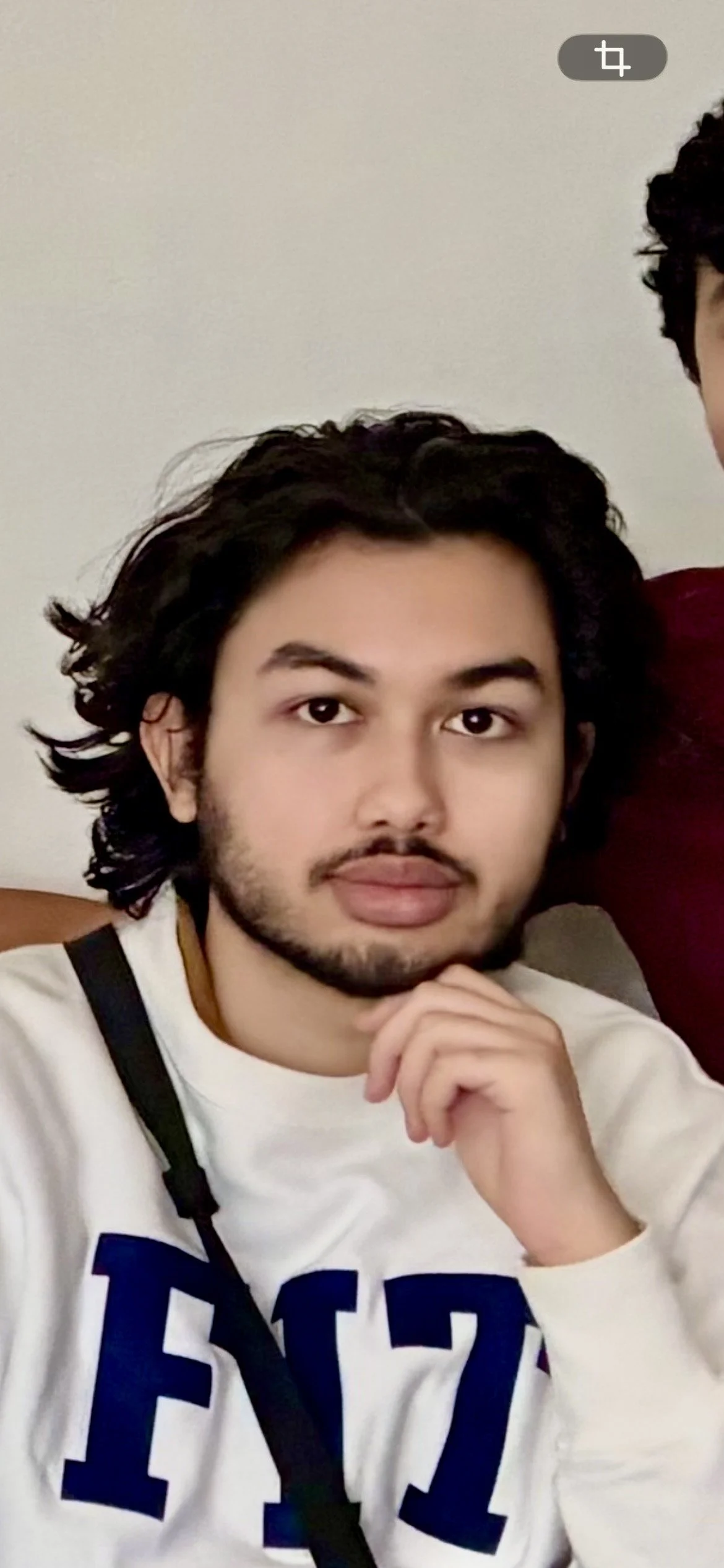 A young man with curly hair and a beard resting his chin on his hand, looking at the camera, wearing a white sweatshirt with large blue letters and a black strap across his chest, sitting against a plain wall.