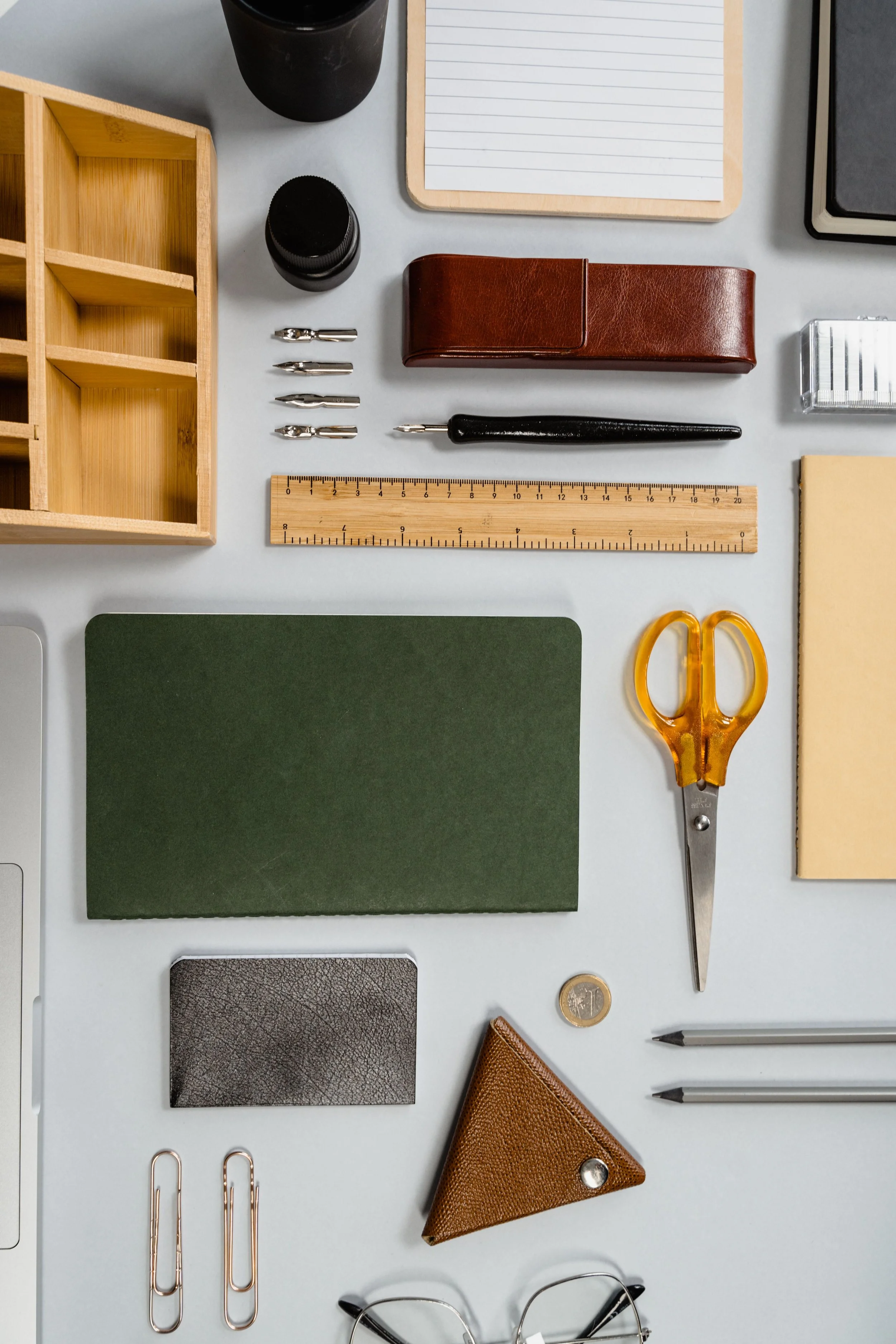 Flat lay assortment of office or stationery items, including a wooden organizer, black cups, lined notepad, red leather case, fountain pen, wooden ruler, green felt pad, textured gray swatch, brown triangular wallet, gold paper clips, gold coin, eyeglasses, two pens, and yellow scissors on a white surface.