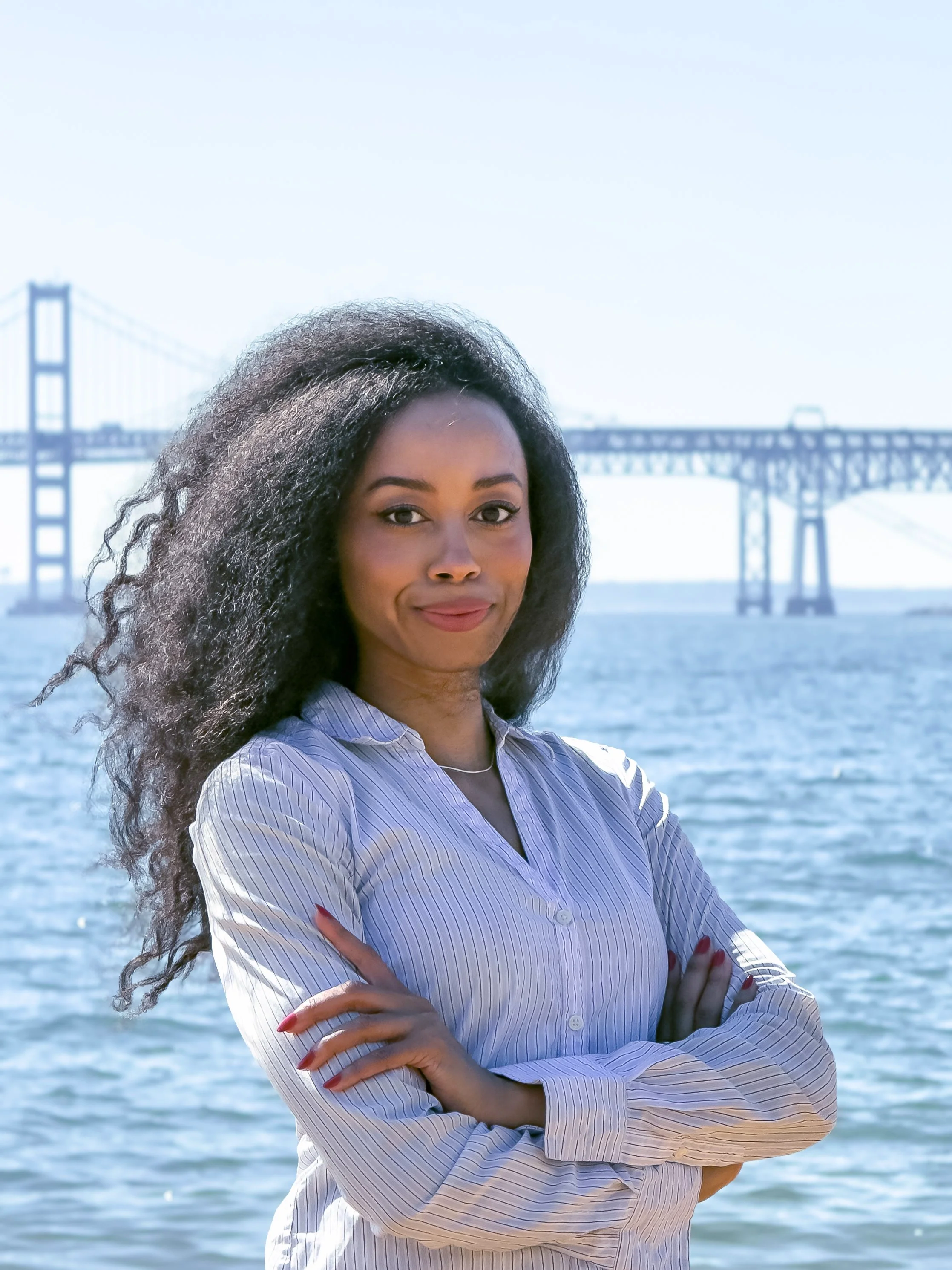 A woman with long curly hair and a striped shirt standing in front of water with a large bridge in the background.