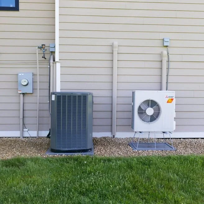Exterior of a building showing HVAC units, pipes, and electrical box against siding, with a grassy area in the foreground.