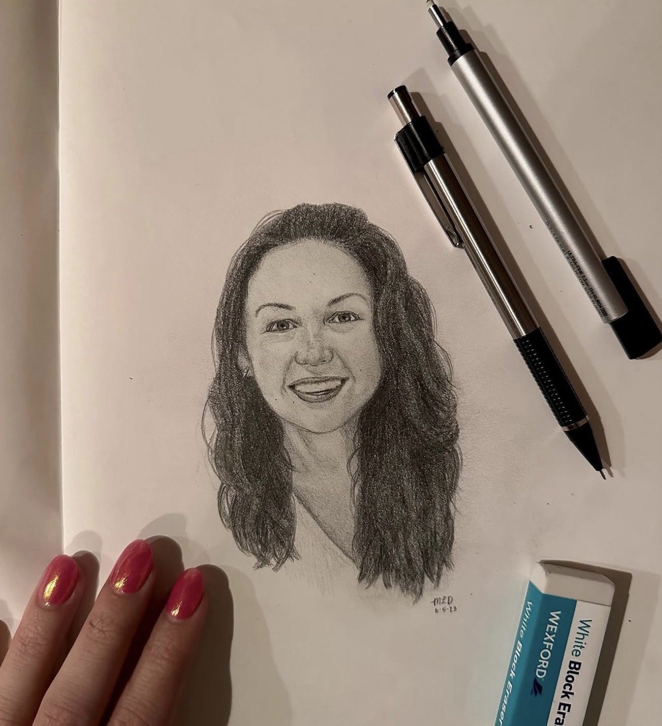 A detailed pencil drawing of a woman with long, curly hair, smiling, on a sheet of paper. Two pens and a tube of white correction fluid are placed on the paper. A person's hand with bright pink painted nails is near the bottom left corner, touching t