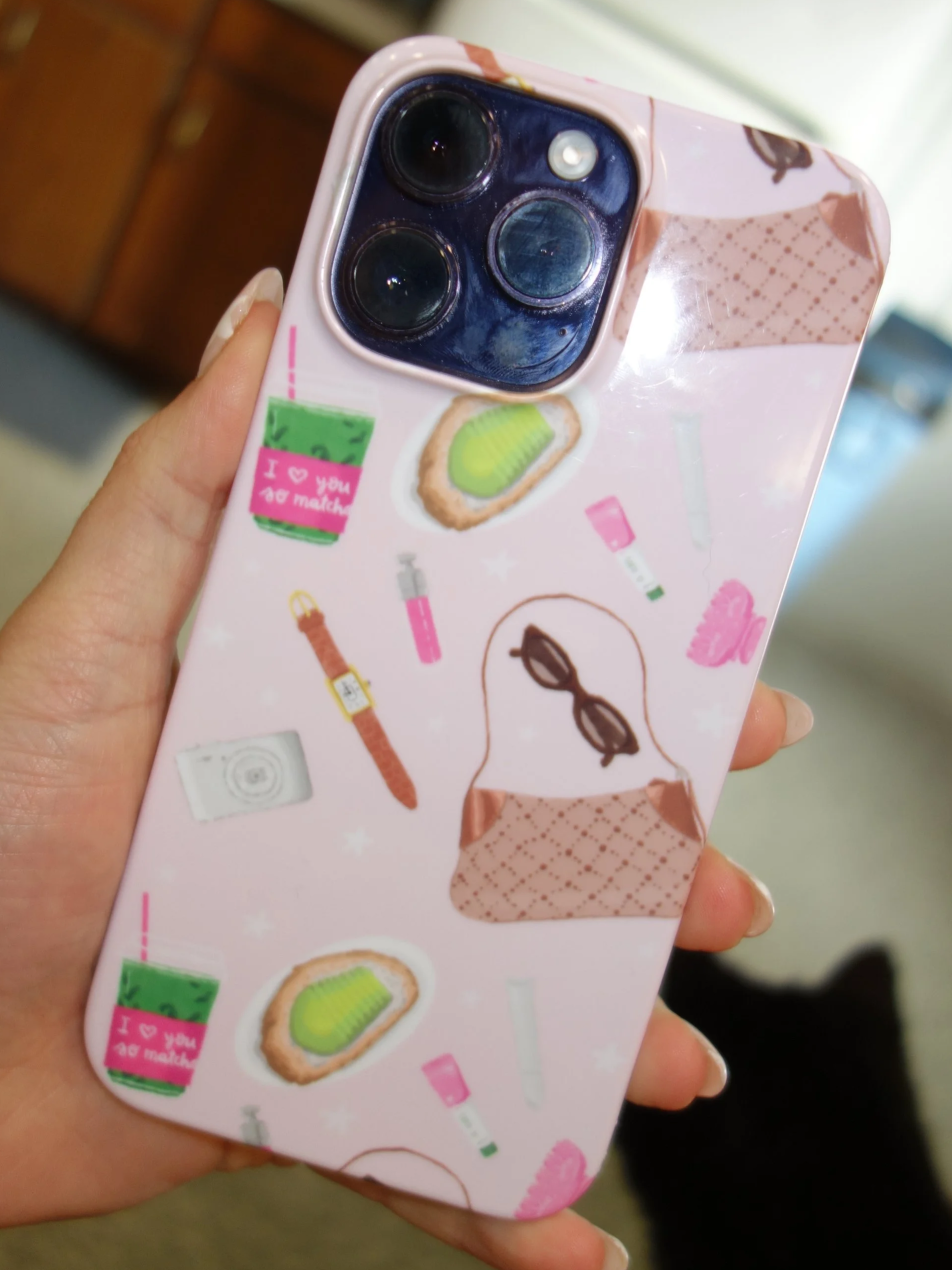 A close-up of a hand holding a phone with a pink case decorated with stickers of various items like a green drink, a slice of toast with avocado, a pair of sunglasses, and beauty products. The phone has a triple camera setup.