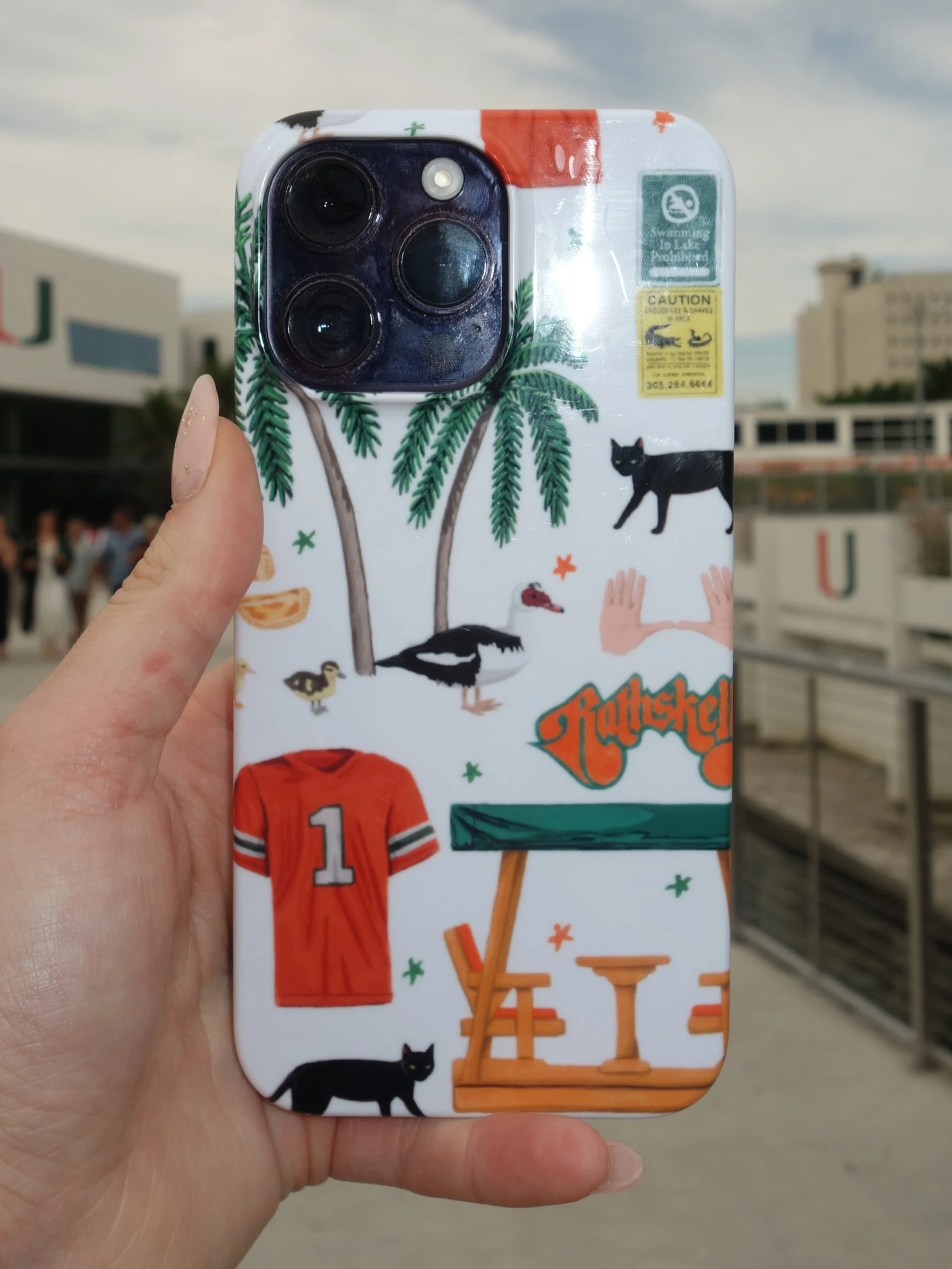 Close-up of a person holding a smartphone with a stickered case, featuring various illustrated objects including a black cat, trees, a wooden bench, a football jersey with the number 1, and a table, with background consisting of buildings and a cloud