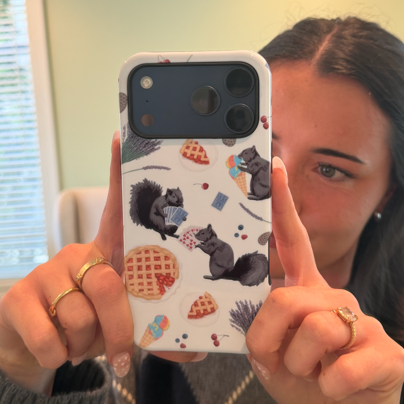 A person taking a selfie with a phone that has a case decorated with cute illustrations of squirrels, acorns, and food items like pie and ice cream cones. The person has dark hair, earrings, and is wearing rings on their fingers.
