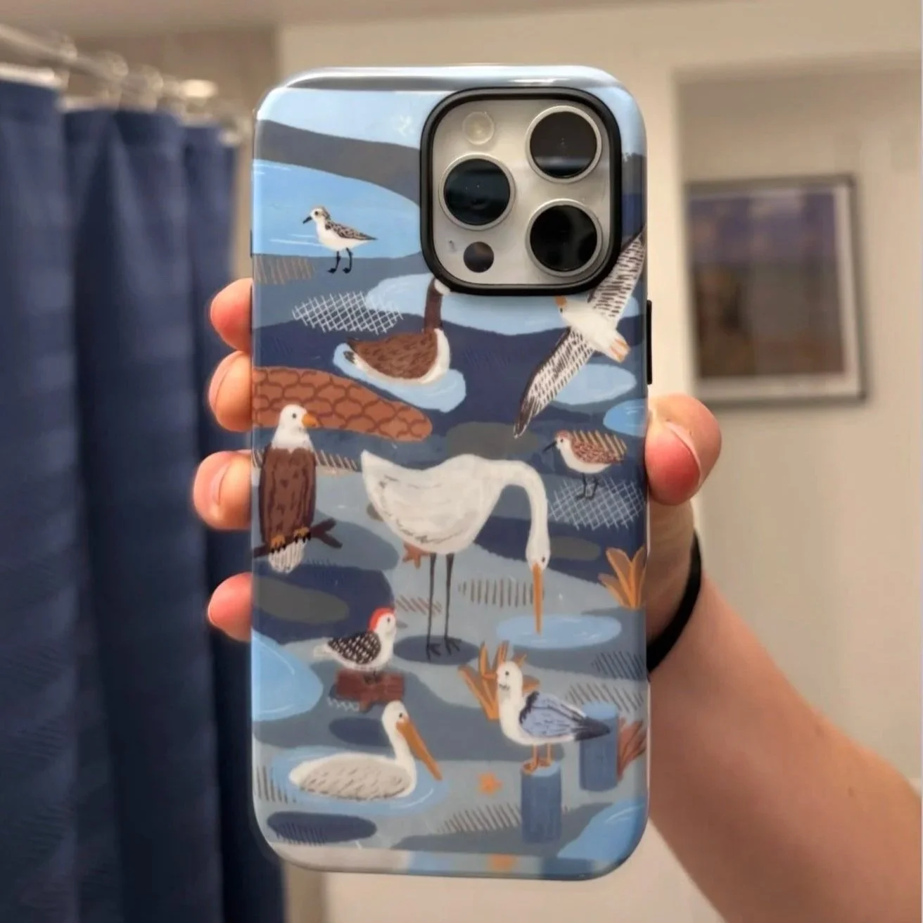 Smartphone with a case decorated with illustrated waterfowl such as herons, ducks, seagulls, and other birds, set against a water and sky background.