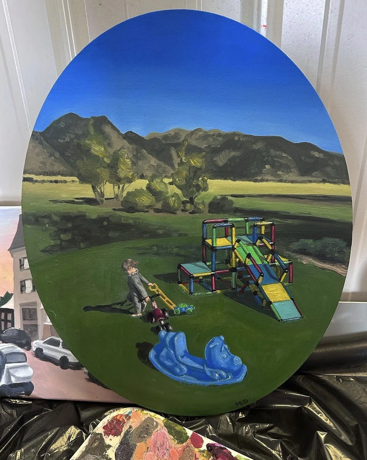 Painting of a landscape with mountains, trees, and a children's playground with slides, a small boy riding a toy mower, and a pond, set against a blue sky with clouds.