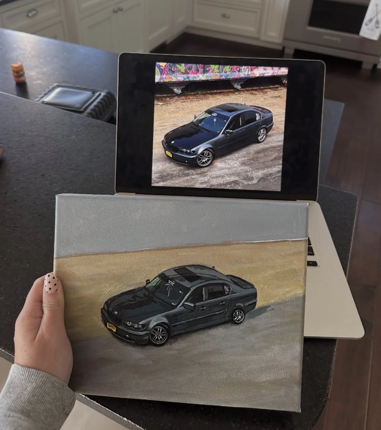 Photograph of a person holding a painting of a black car, with a tablet displaying the same car above it. The scene is set on a kitchen countertop.