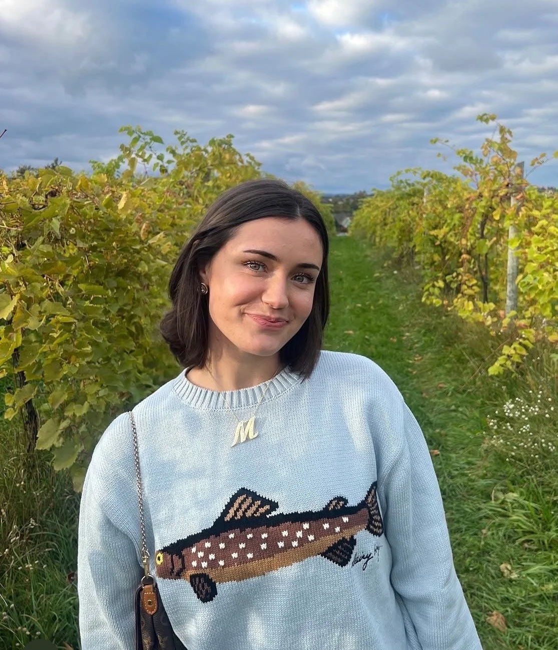 A young woman with dark hair and light skin standing in a vineyard, wearing a light blue sweater with a fish design and a gold letter 'M' necklace, smiling at the camera with a cloudy sky overhead.