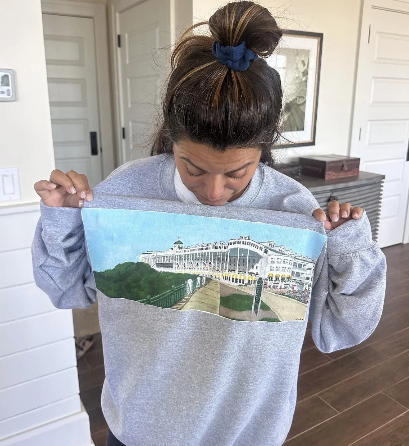 A woman with light brown hair tied up with a blue scrunchie is looking down at a colorful picture she is holding in front of her. She is wearing a gray sweatshirt and is indoors with white walls, a framed photograph, and a side table in the backgroun