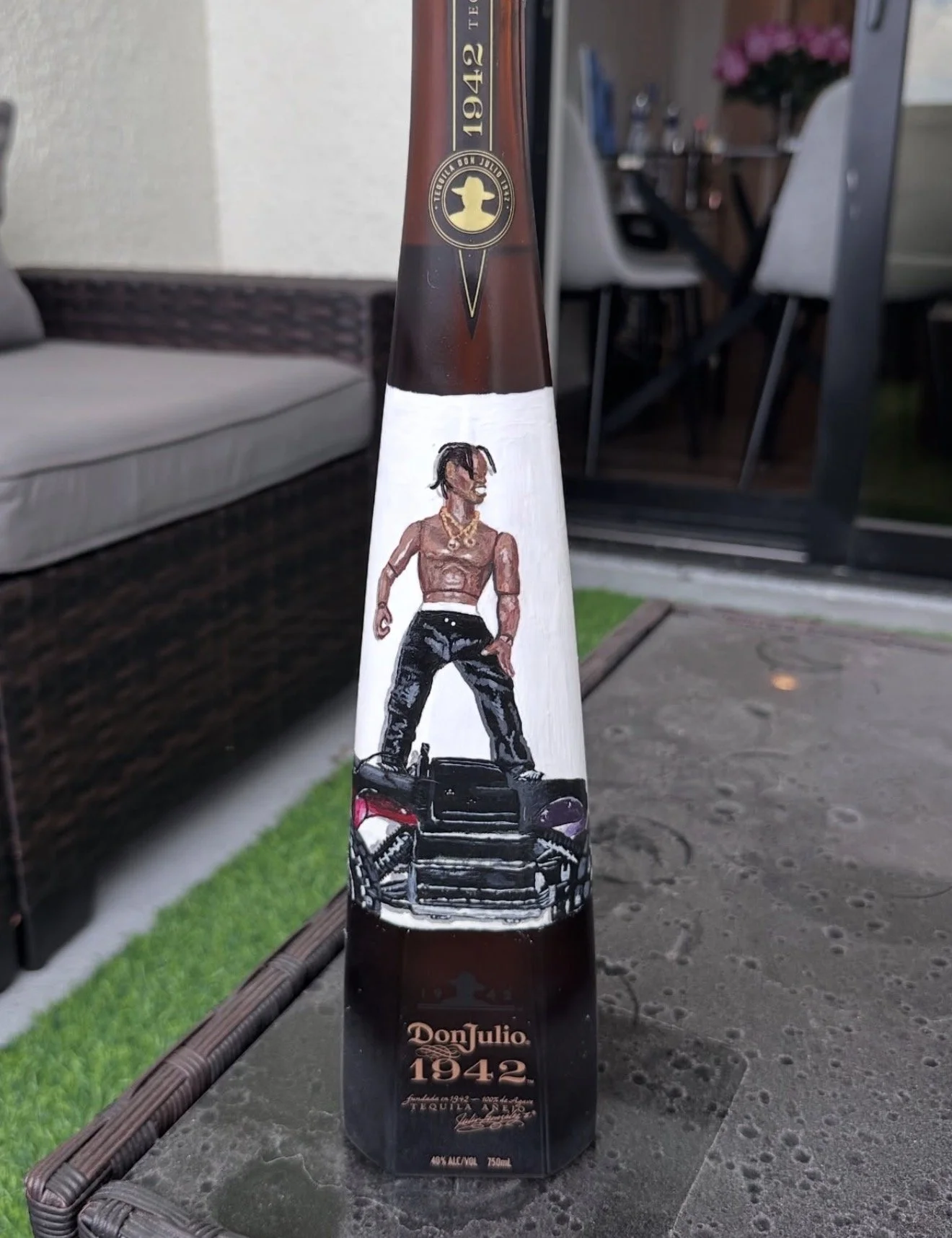 A bottle of Don Julio 1942 tequila with a custom label featuring a muscular animated character with dreadlocks, standing on a car. The background shows an outdoor patio with a sofa, chairs, a table with flowers, and an open door.