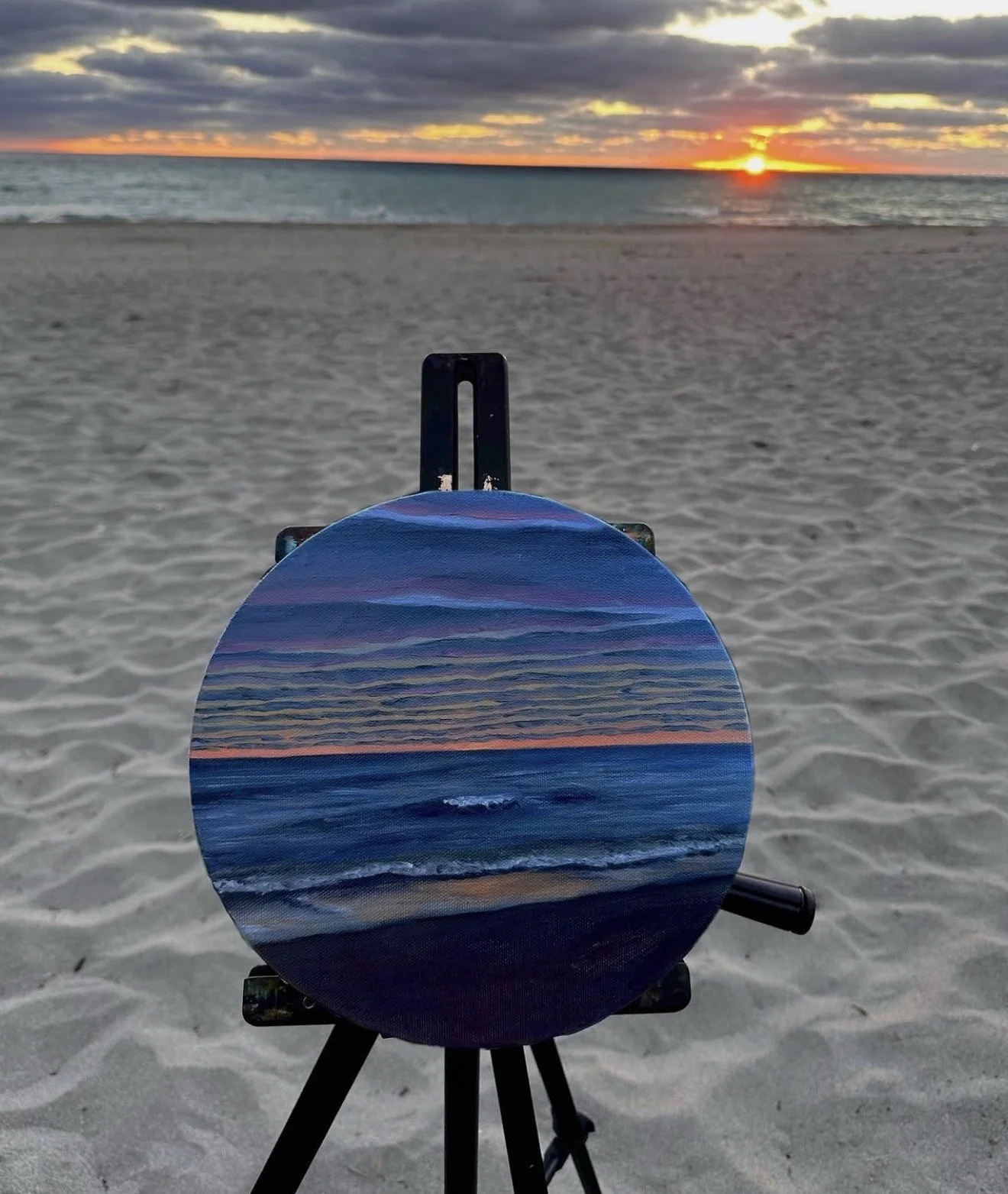 Painted artwork of a beach scene with the ocean, sky, and sunset, set on a circular canvas mounted on an easel at the beach with sand and sea in the background.