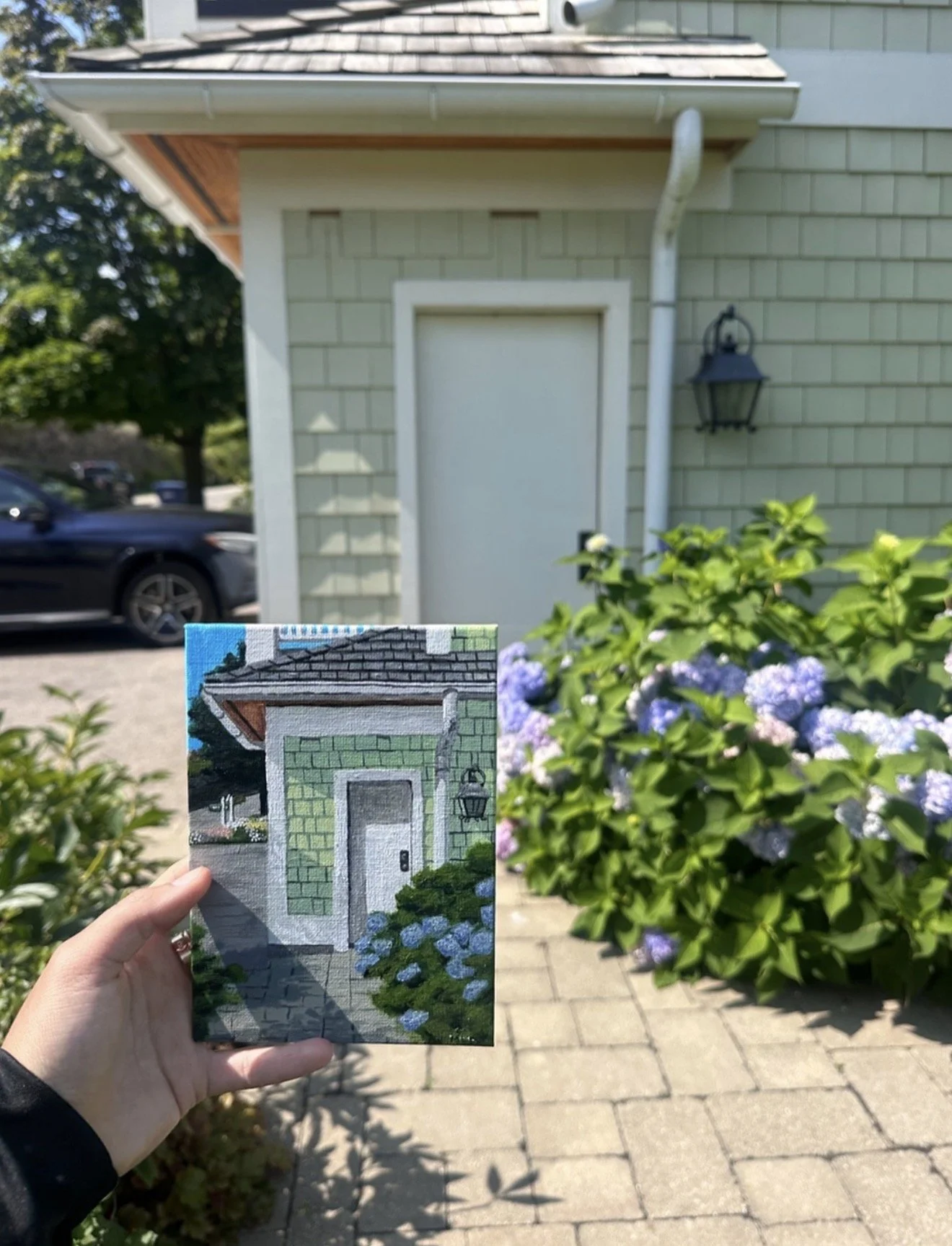 A hand holding a small painting or print of a house facade with green siding and a door, with purple flowers at the bottom, in front of the real house which has green siding, a white door, and a lantern. There are bushes with purple flowers in the fo