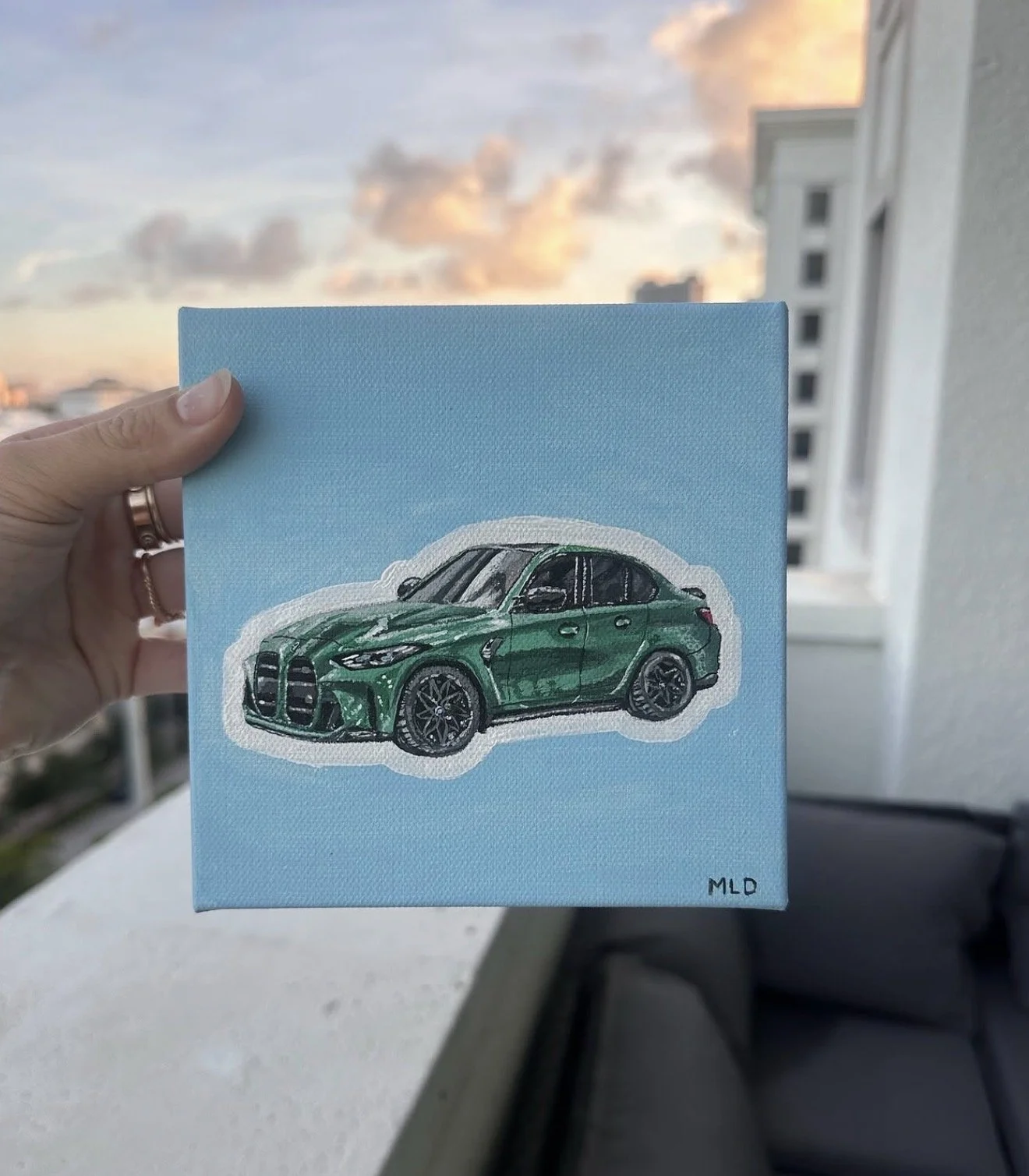 Photo of a person holding a canvas with a painted green sports car, with a cloudy sky in the background.