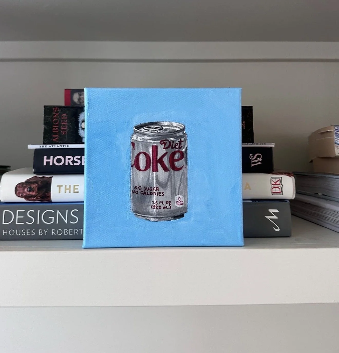 Small canvas painting of a Diet Coke can with the label 'No Sugar No Calories' on a blue background, placed on a shelf among books.