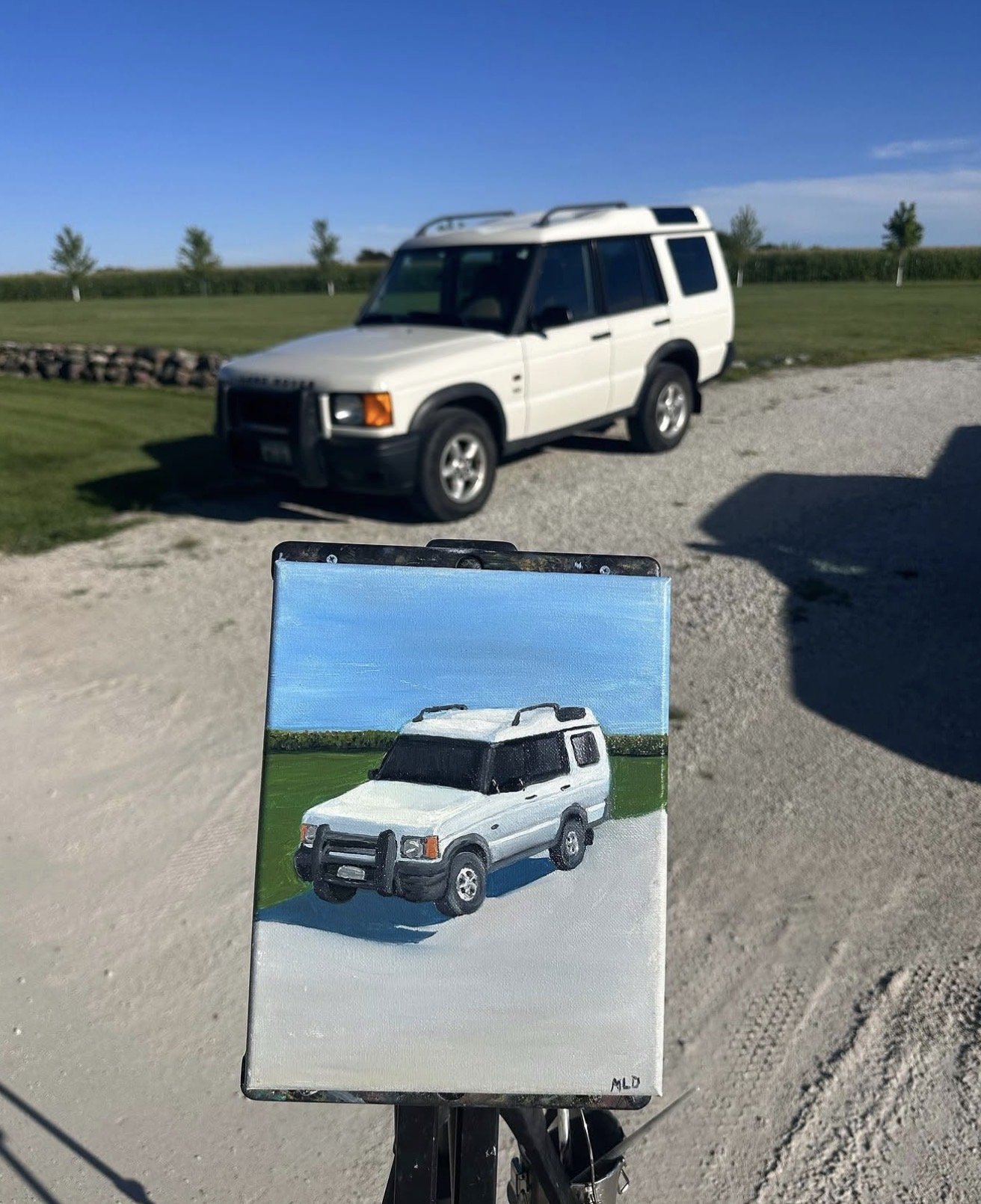 A landscape painting of a white SUV on a grassy field with a bright blue sky, held in front of an actual white SUV parked on a gravel surface with green grass and trees in the background.