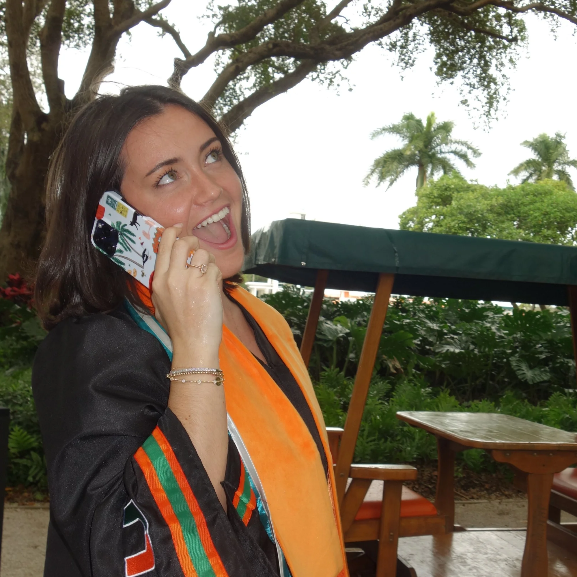 A woman with dark hair smiling and talking on a cellphone, outdoors with trees and greenery in the background, wearing a colorful scarf and black jacket.