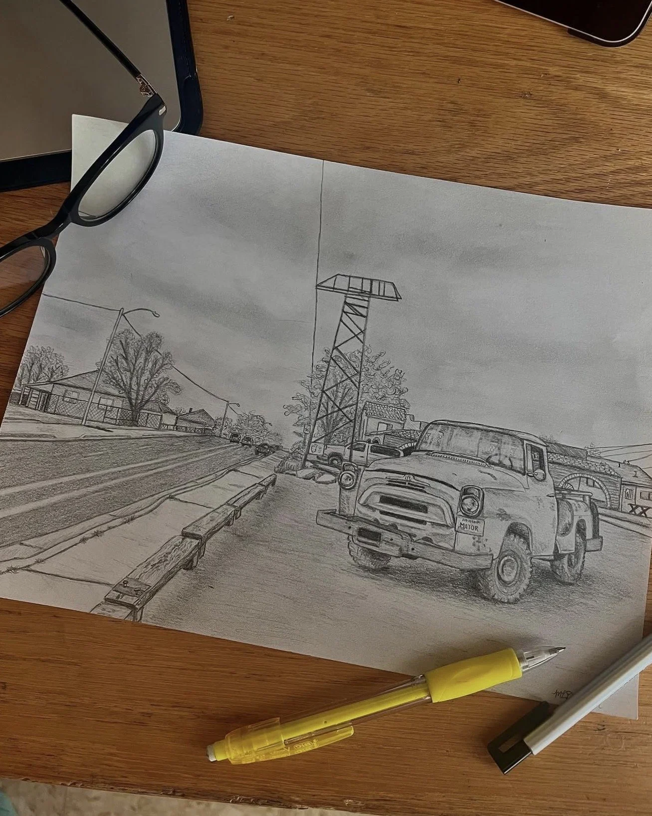 Black and white sketch of a city street scene on a piece of paper, featuring a vintage truck in the foreground, utility pole, streetlights, trees, houses, and cars on the road, with a pen placed on the wooden table.
