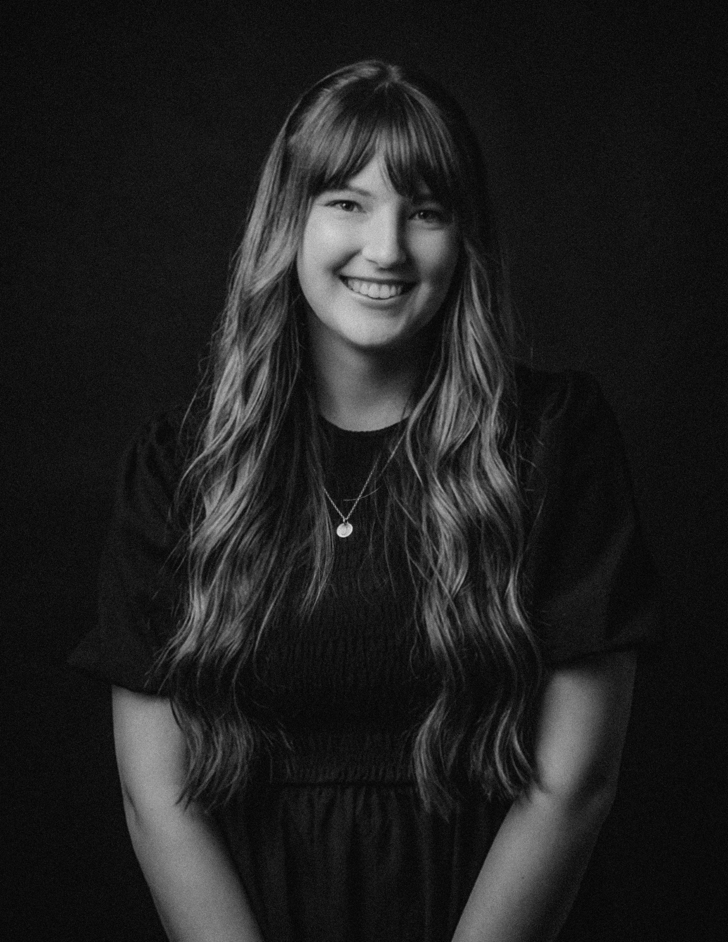 A woman with long wavy hair smiling, wearing a dark top and a necklace, against a dark background.