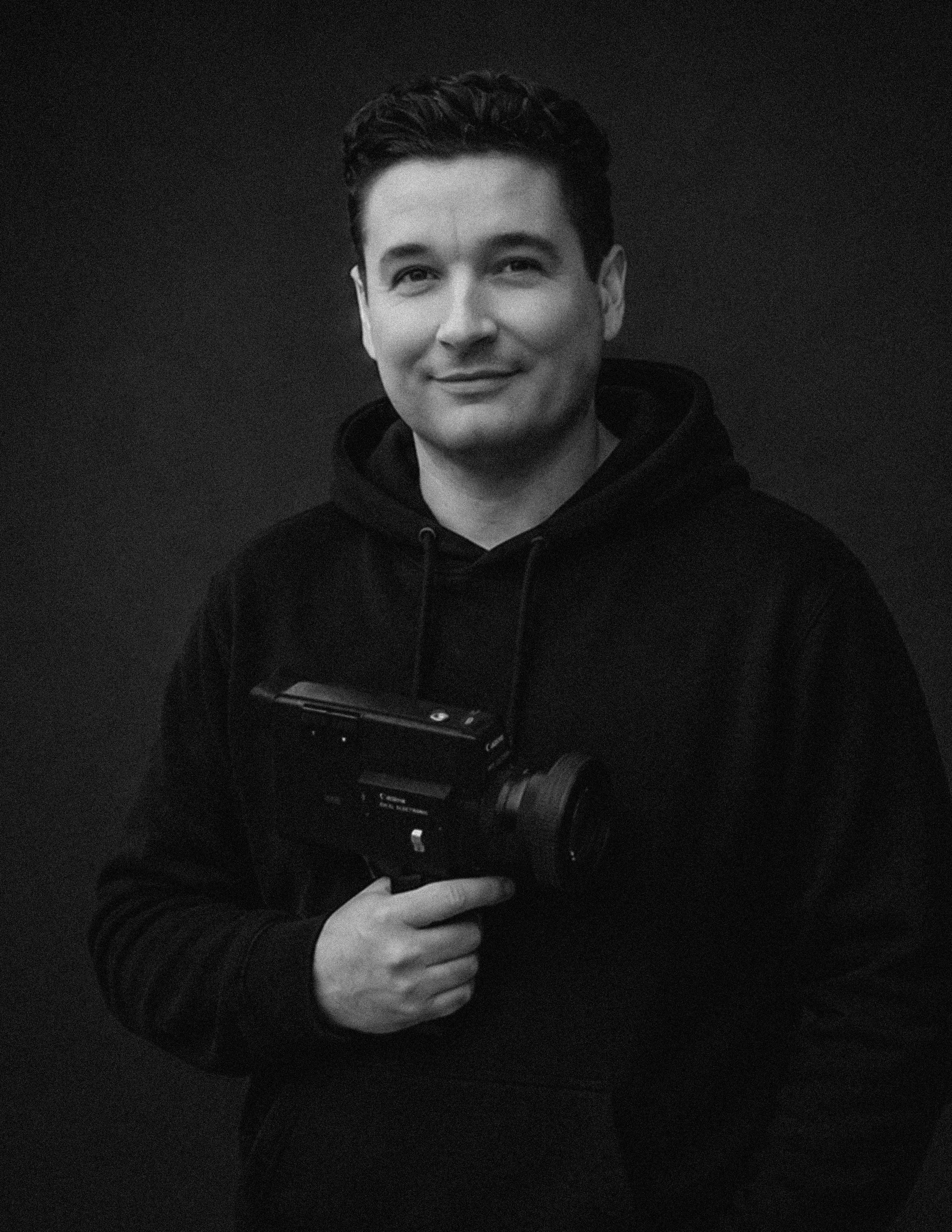 Black and white photo of a young man with dark, curly hair, wearing a hoodie and smiling slightly while holding a camera in front of his chest.