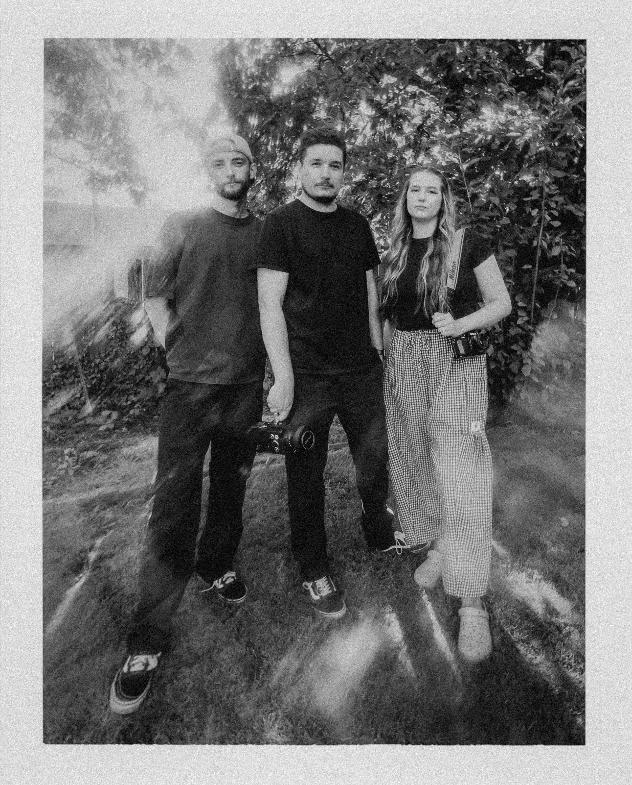 Three young adults standing outdoors in front of leafy trees, with a film camera hanging from the woman’s shoulder. The photo is in black and white.