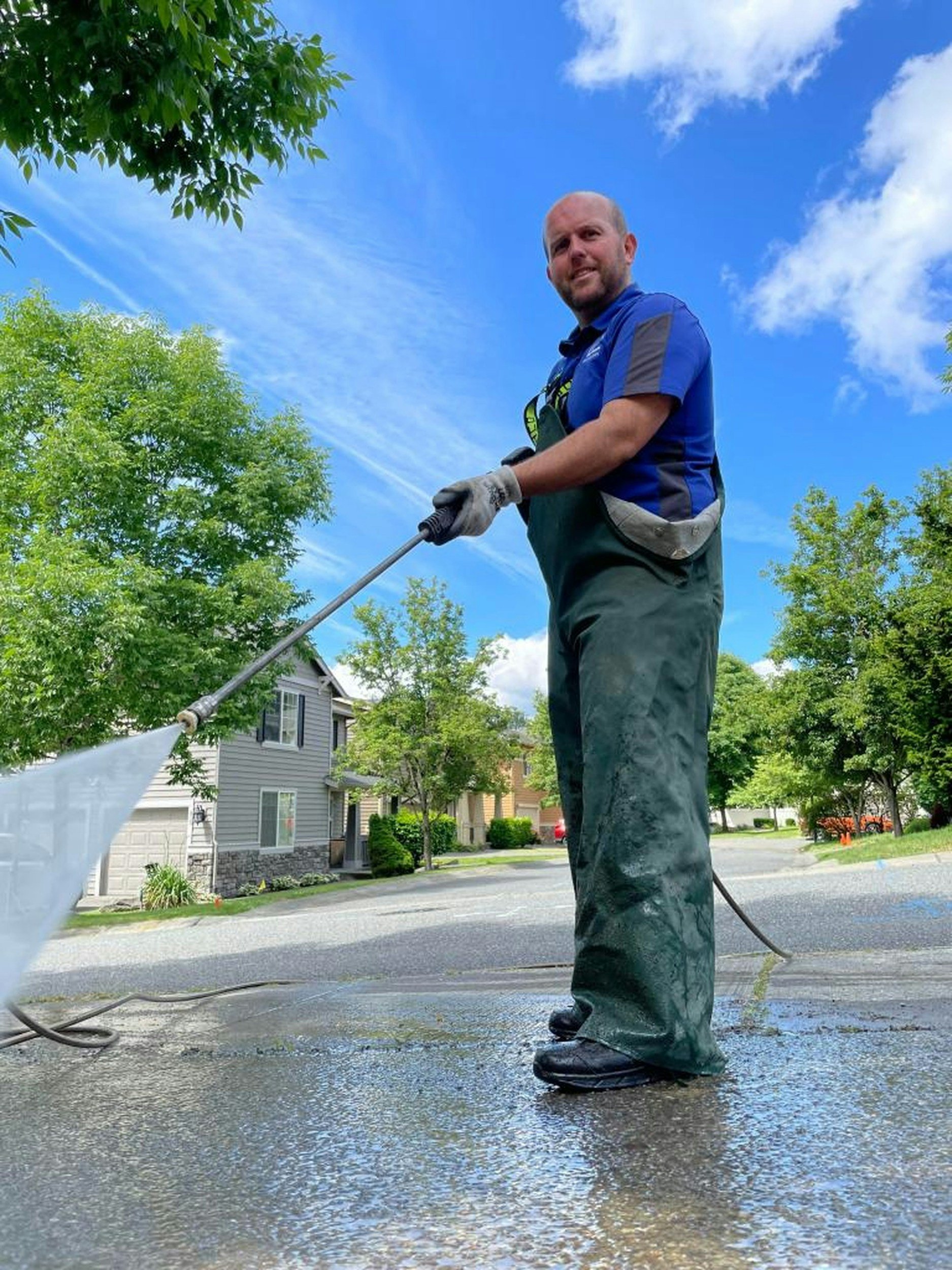 A man wearing waterproof pants and gloves is cleaning a driveway with a pressure washer on a sunny day, with trees and houses in the background.