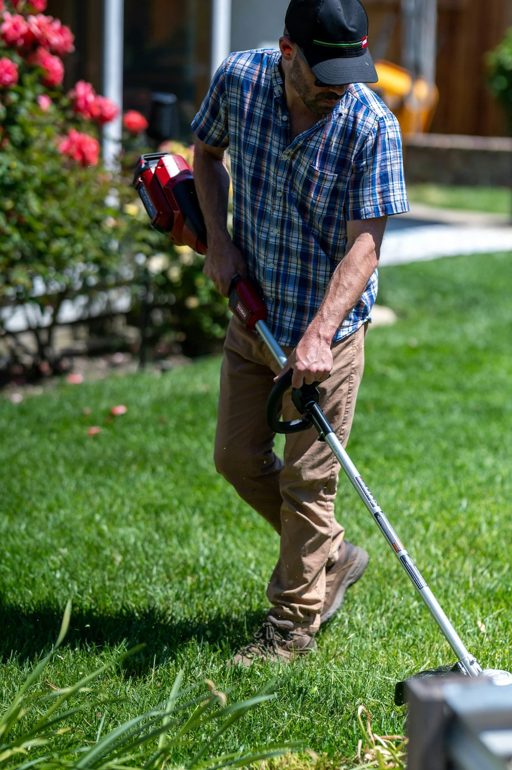 A man wearing a black cap, glasses, and a plaid shirt is using a weed trimmer on a lush green lawn. He is standing and bent slightly forward, focused on trimming the grass near some flowers.