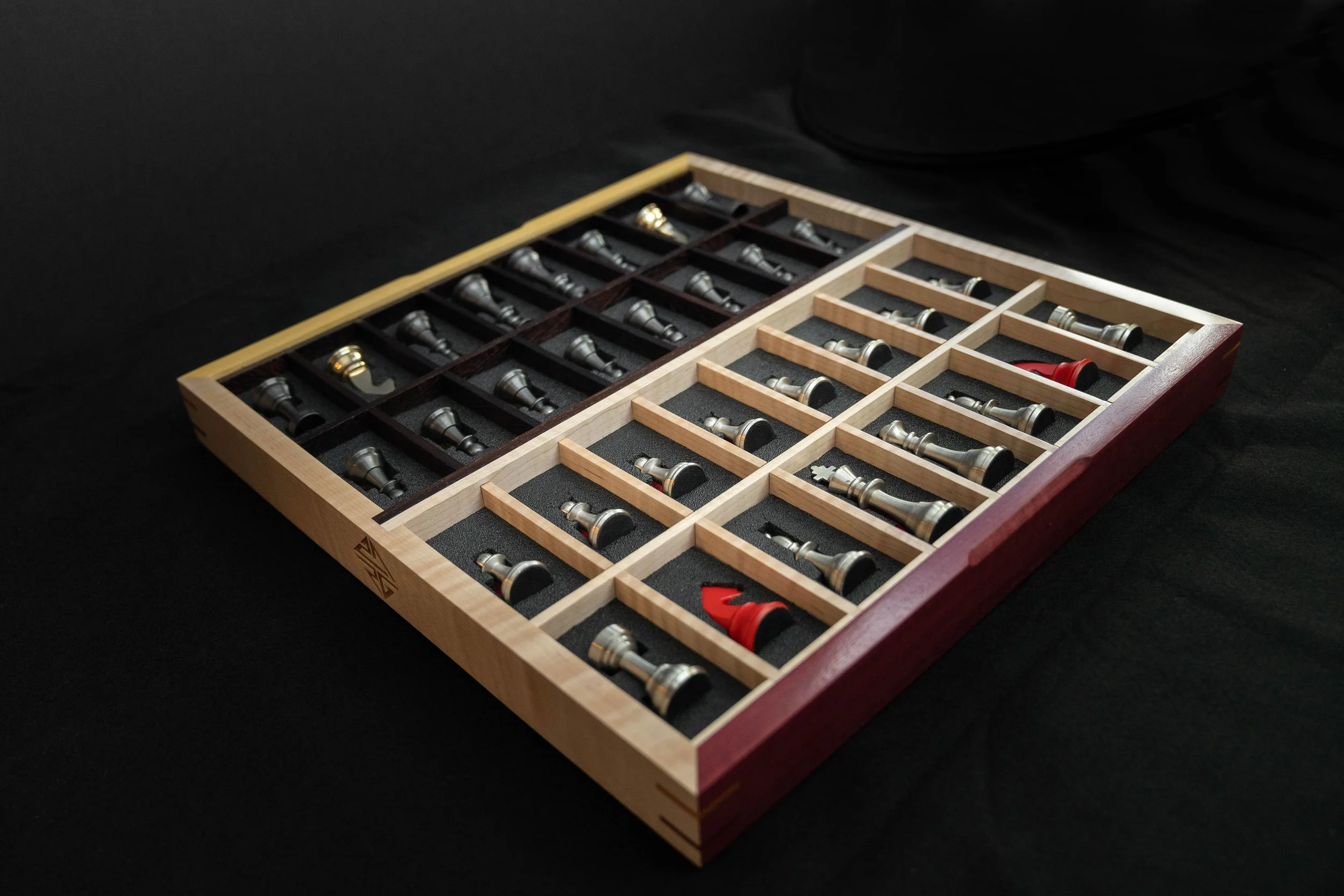 A chess set with black and silver pieces in a wooden box with compartments, on a black surface.