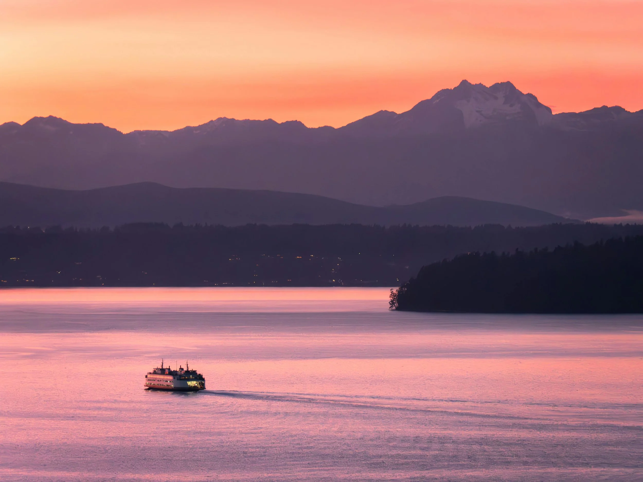 A passenger ferry boat traveling across a calm body of water during sunset, with mountains in the background and a colorful pink and orange sky.